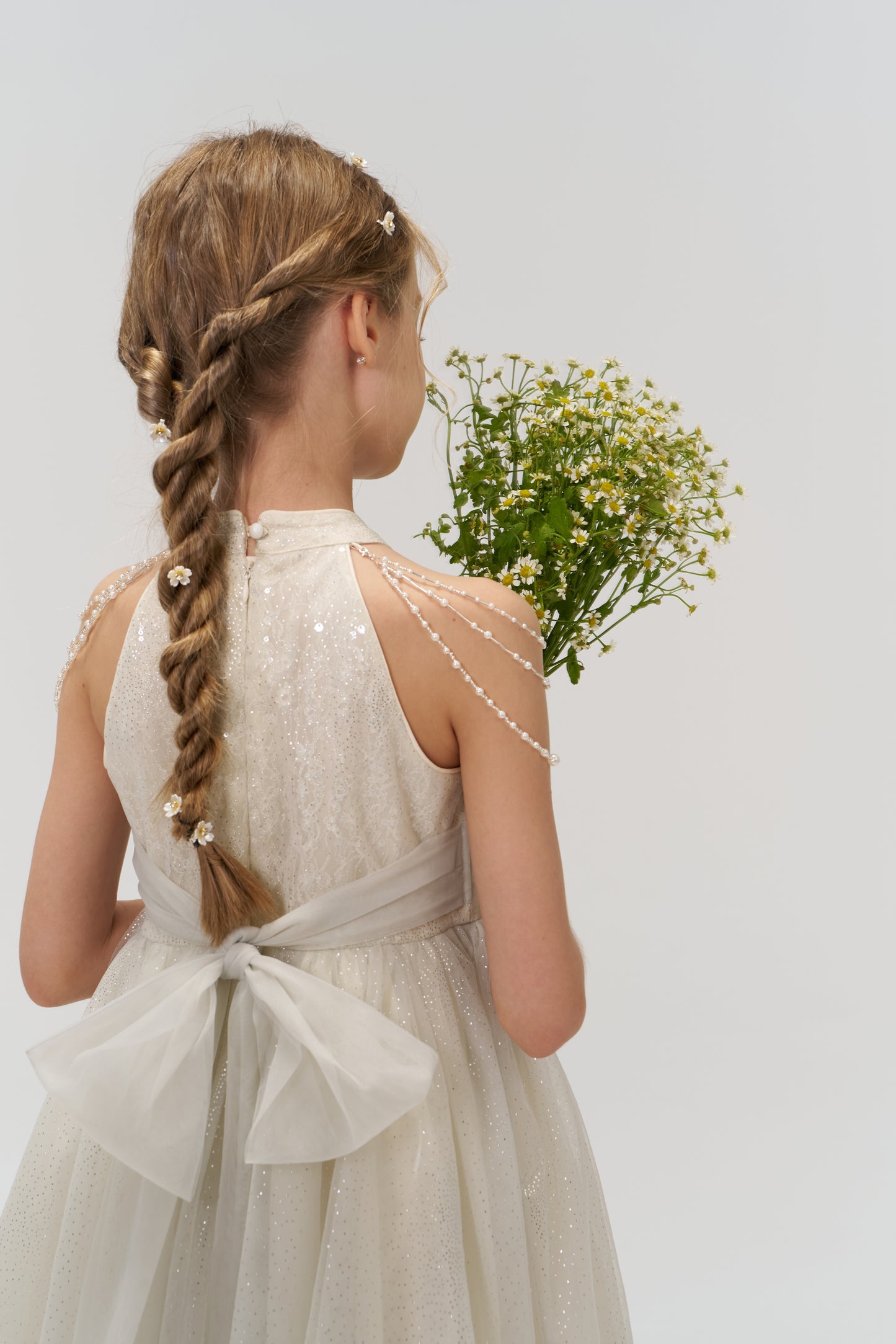 A young girl with a braided hairstyle and small flowers holds yellow wildflowers, wearing KIKI and JOJO's Gilded Whisper Dress—a light halter-neck sequin dress with a large bow at the back.