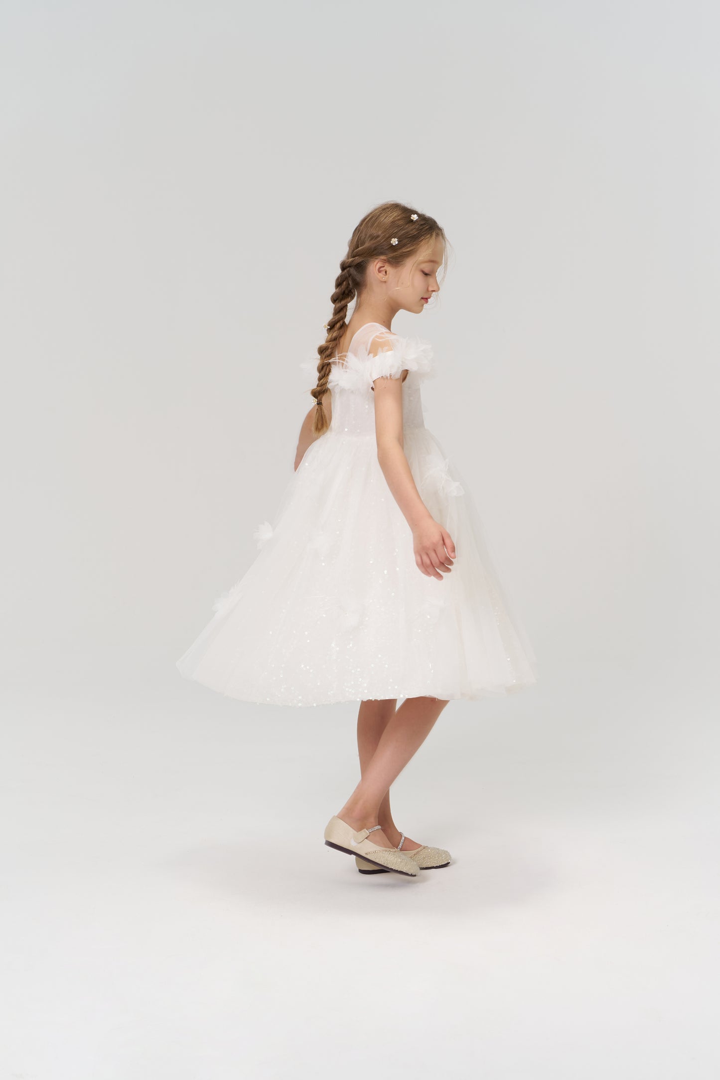 A young girl with braided hair radiates elegance in profile, wearing the KIKI and JOJO Feathered Whisper Dress in blue against a simple light background, paired with light shoes.