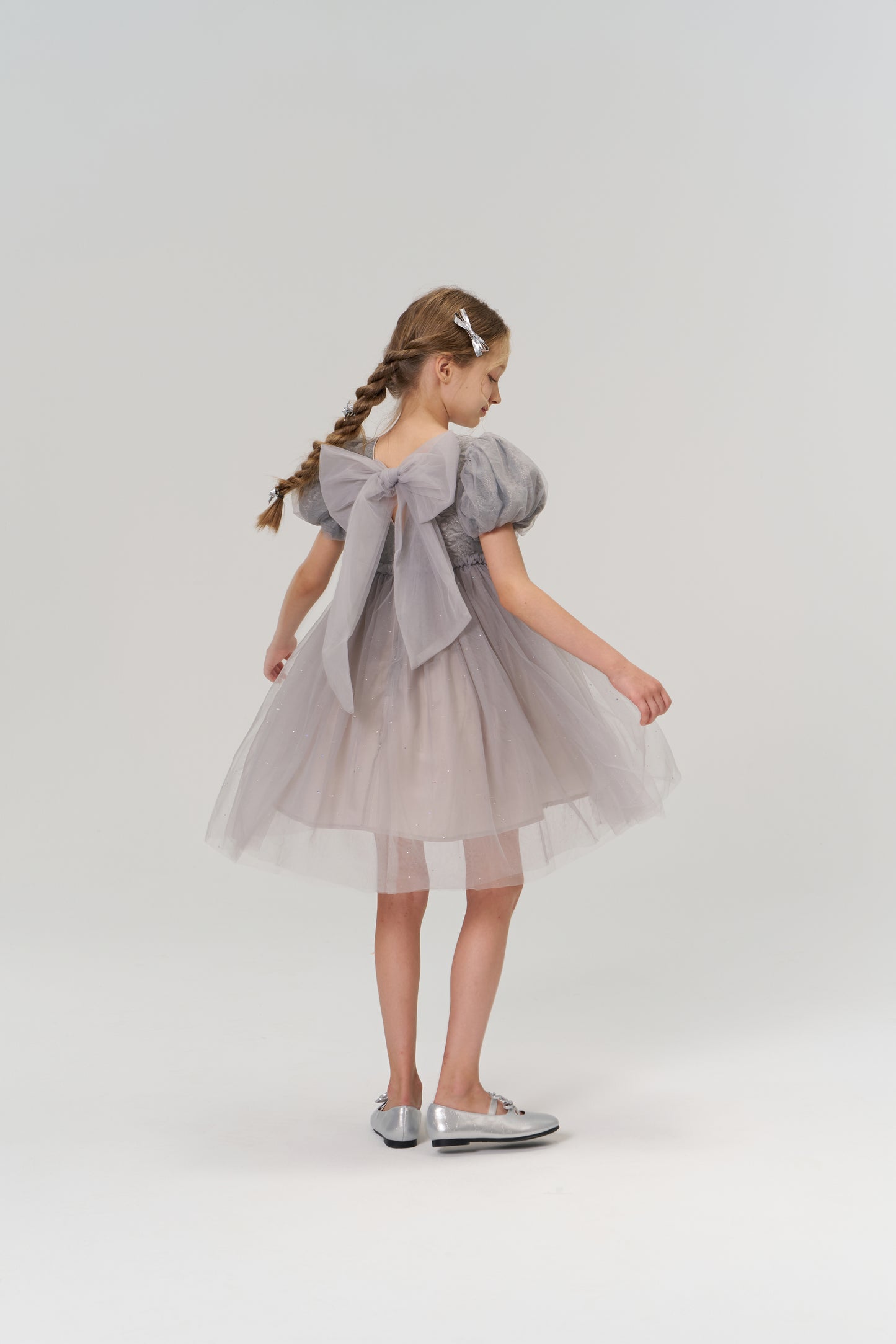 A young girl with braided hair styled with KIKI and JOJO Girls Silver Bow Hair Clips (Set of 2) wears a gray tulle dress with a large back bow and silver shoes, standing against a plain light background.