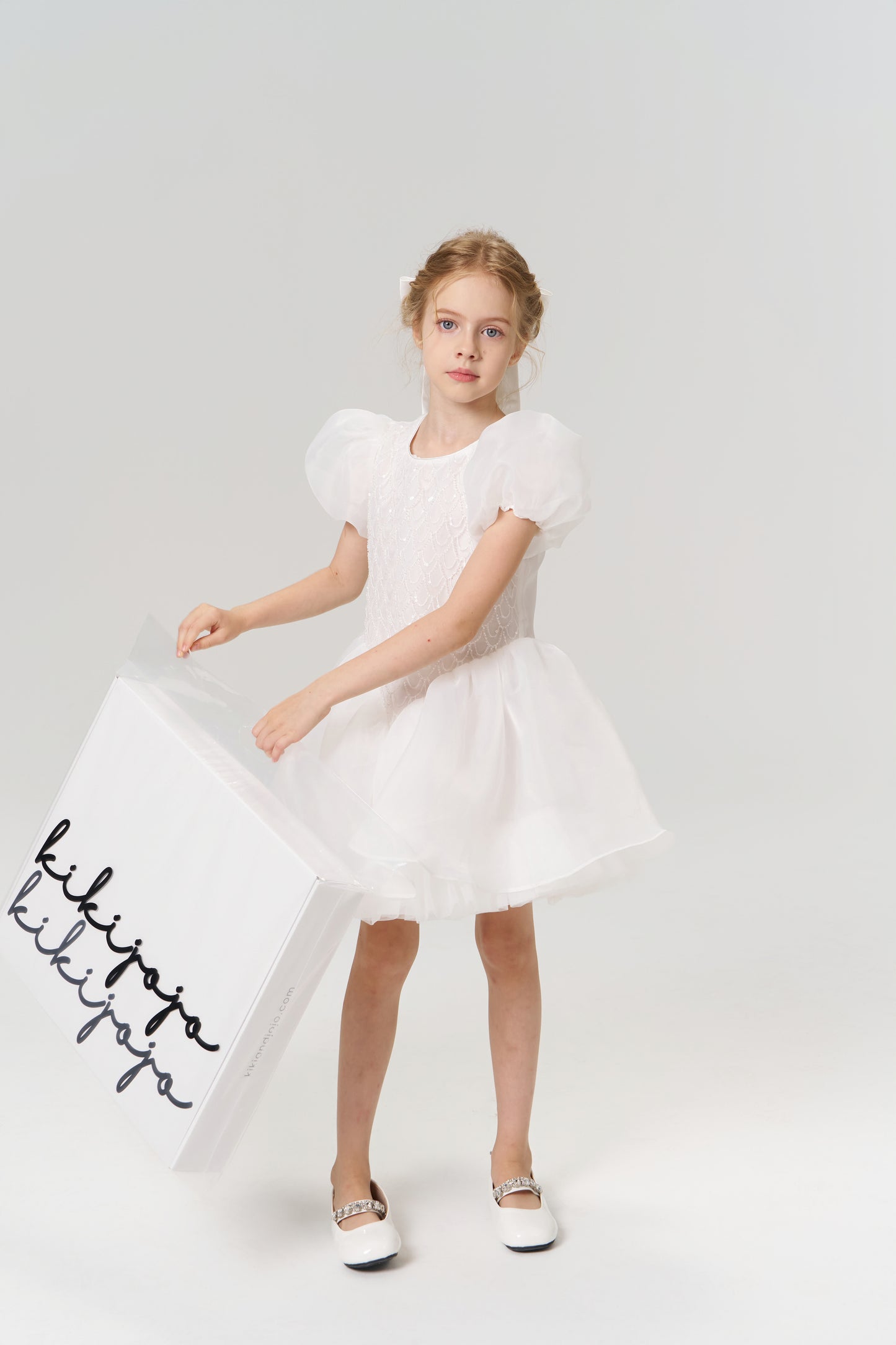 A young girl in a white dress and shoes holds a large KIKI and JOJO Gift Packaging box with elegant black cursive, making it the perfect gift wrap for any special occasion, set against a plain light background.