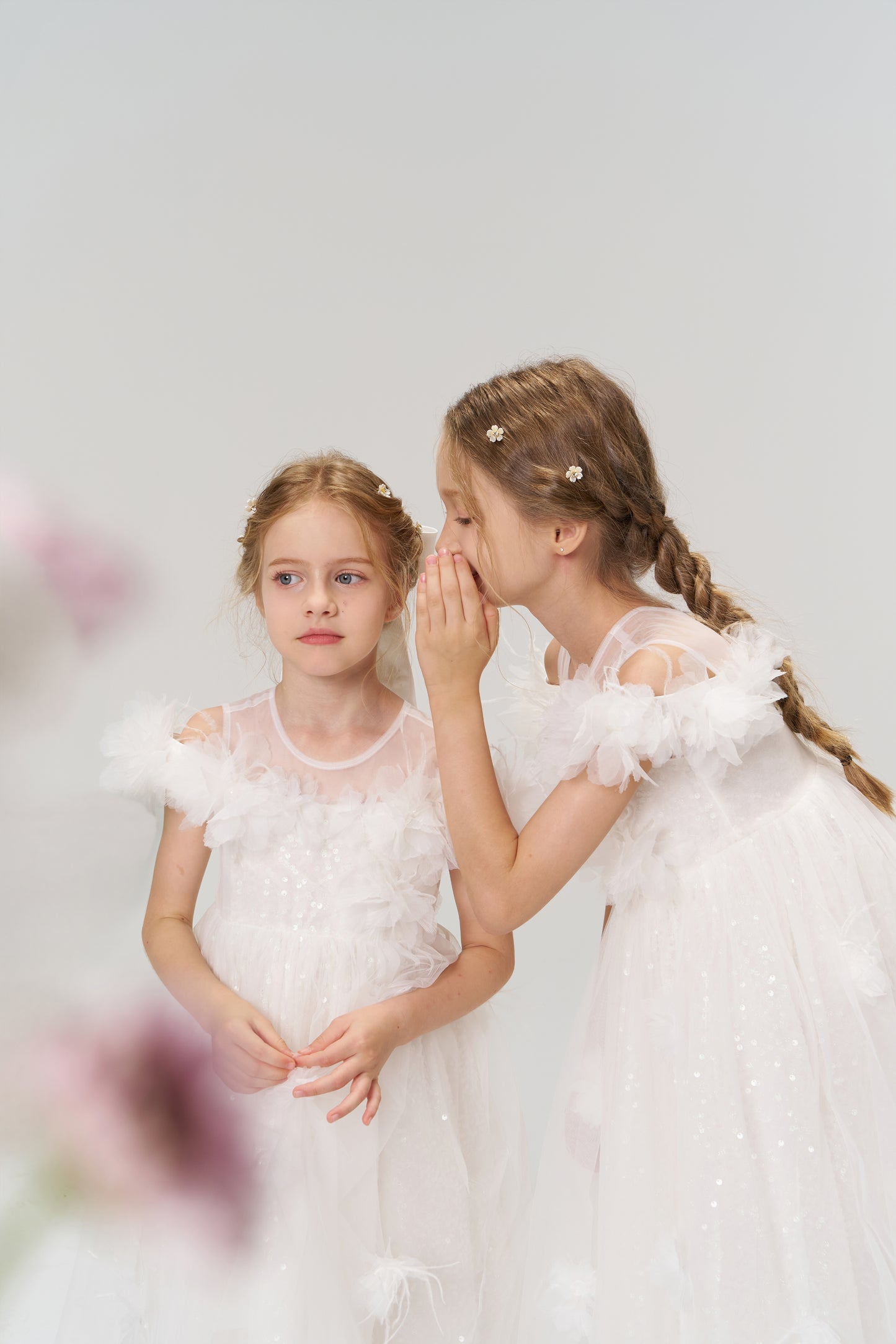 Two young girls wear KIKI and JOJO’s Feathered Whisper Dress, featuring white satin and floral details. Both have braided hair and neutral expressions; one whispers to the other against a light background.