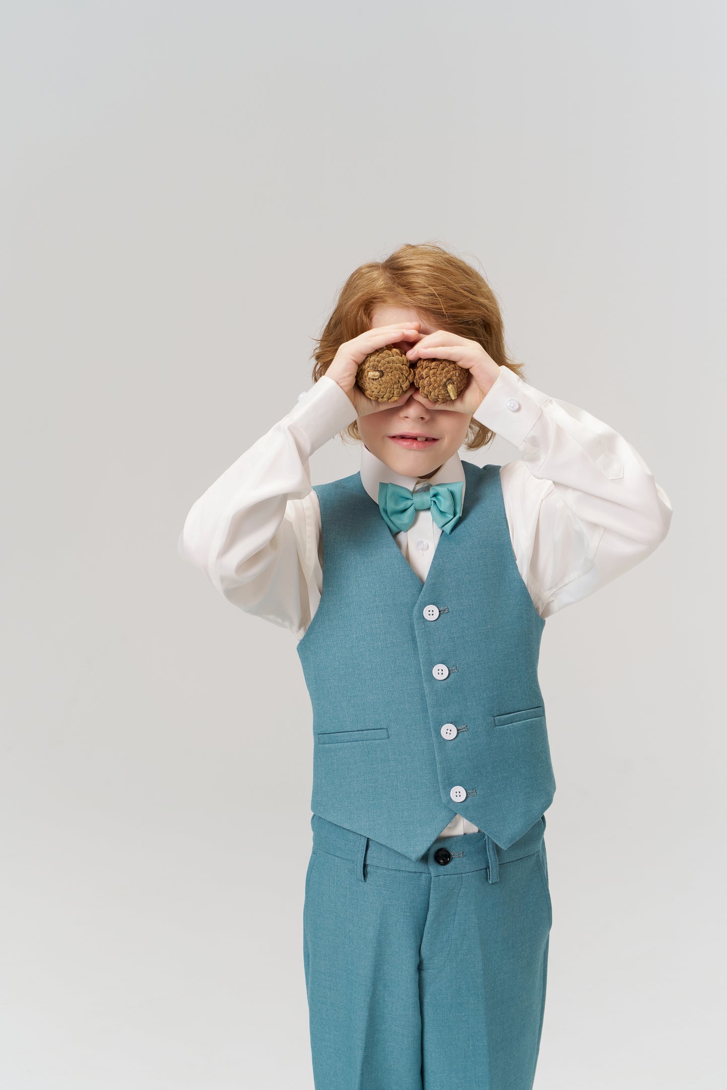 A young boy wears KIKI and JOJO’s Celeste Whimsy Suit Pants with an adjustable waistband and bow tie, playfully holding two cookies to his eyes like binoculars against a plain light background.