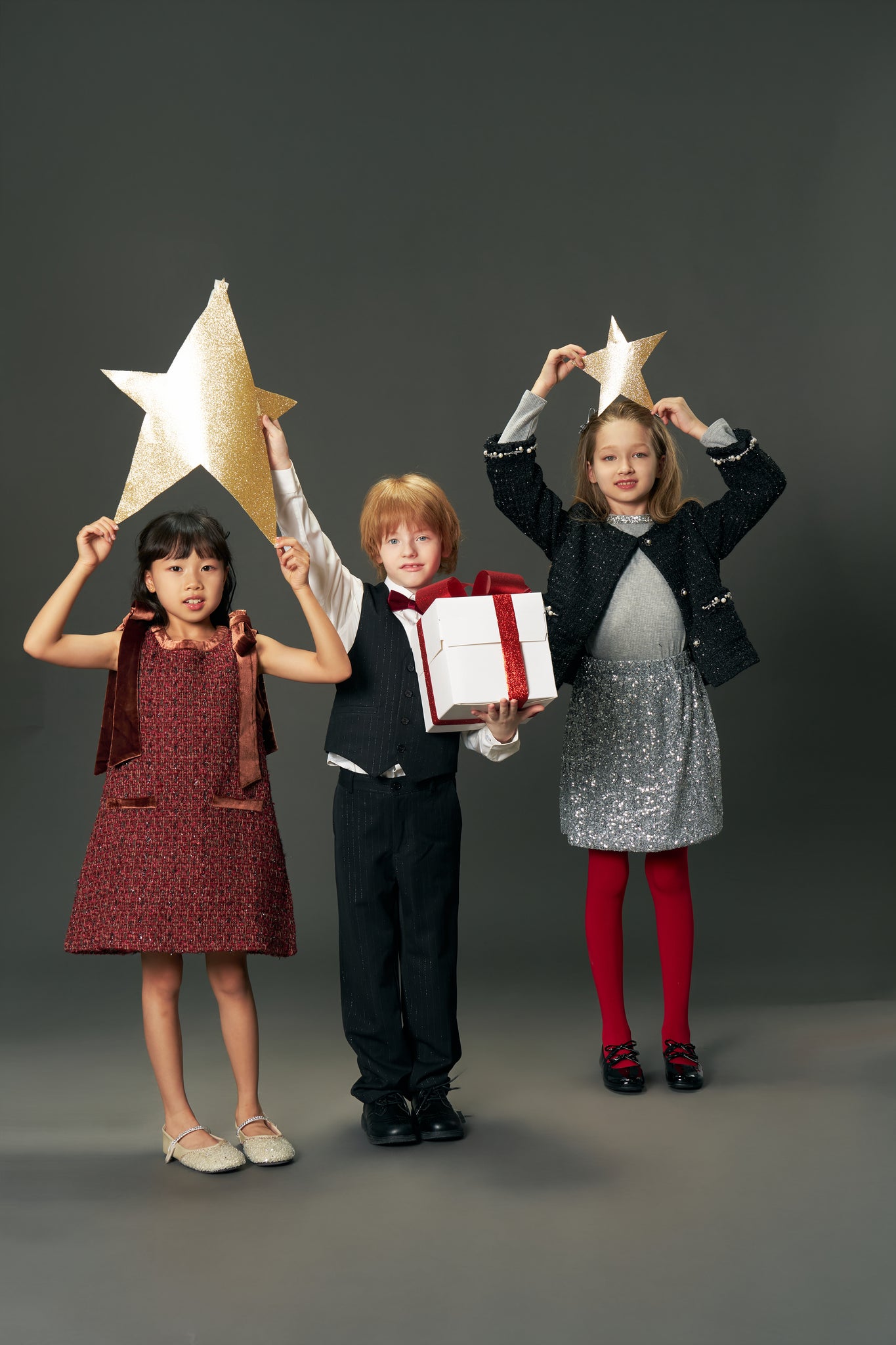 Three children pose against a gray backdrop; one wears the KIKI and JOJO Girls Handmade Tweed Dress with a Burgundy Velvet Bow, while two hold gold star props. The center child in this dress holds a white gift box with a red ribbon.