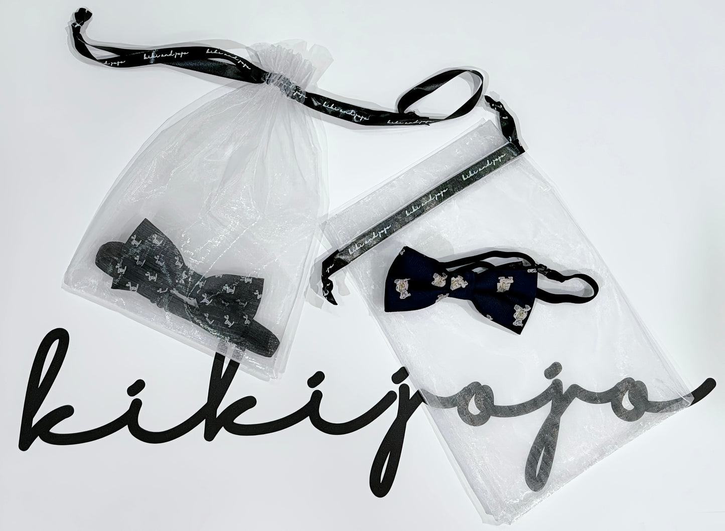 A burgundy satin kids bow tie with bear embroidery from KIKI and JOJO is displayed in a clear drawstring bag above the brand’s name in black script, adding a charming touch.