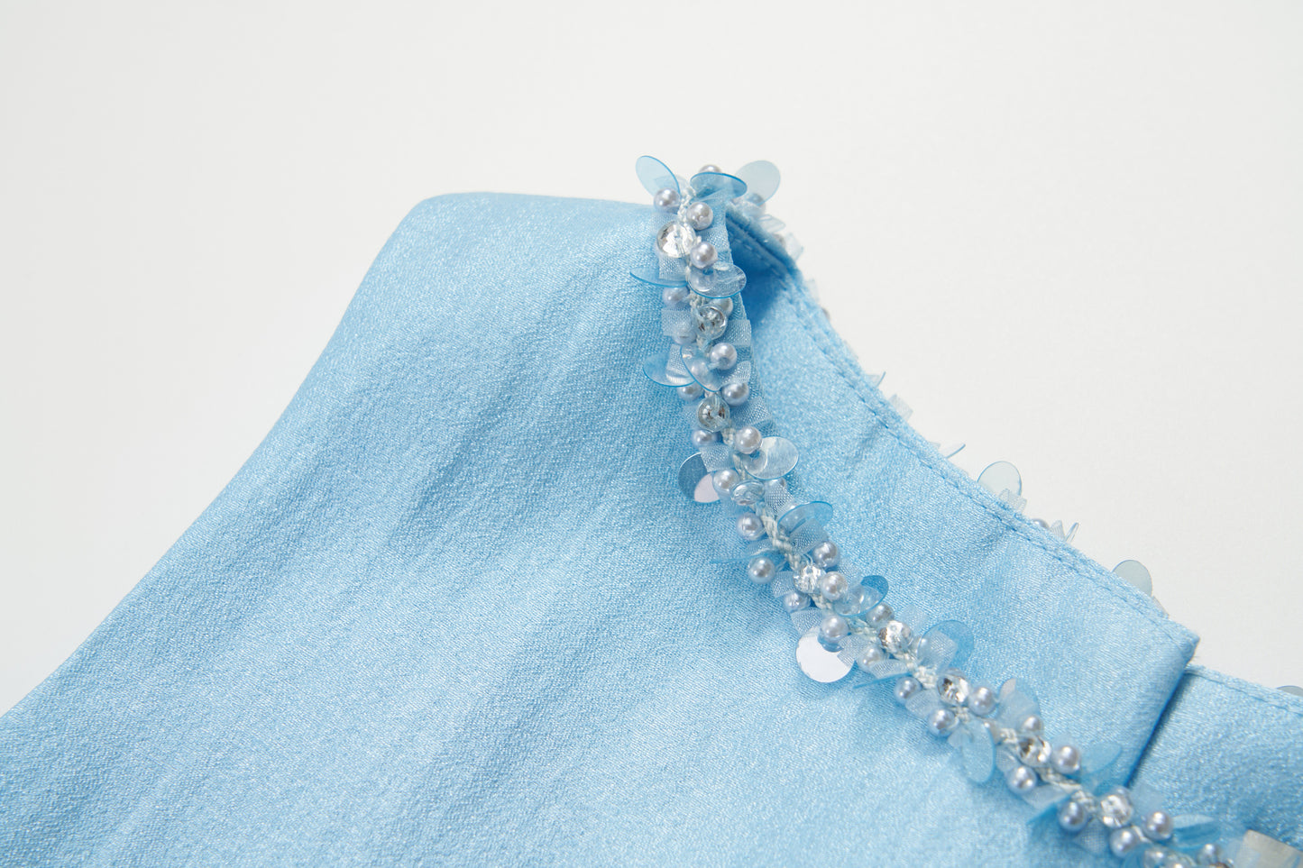 Close-up of the Radiance Satin Crop Top by KIKI and JOJO, crafted from light blue liquid-sheen satin with a hand-beaded pearl and translucent sequin neckline.