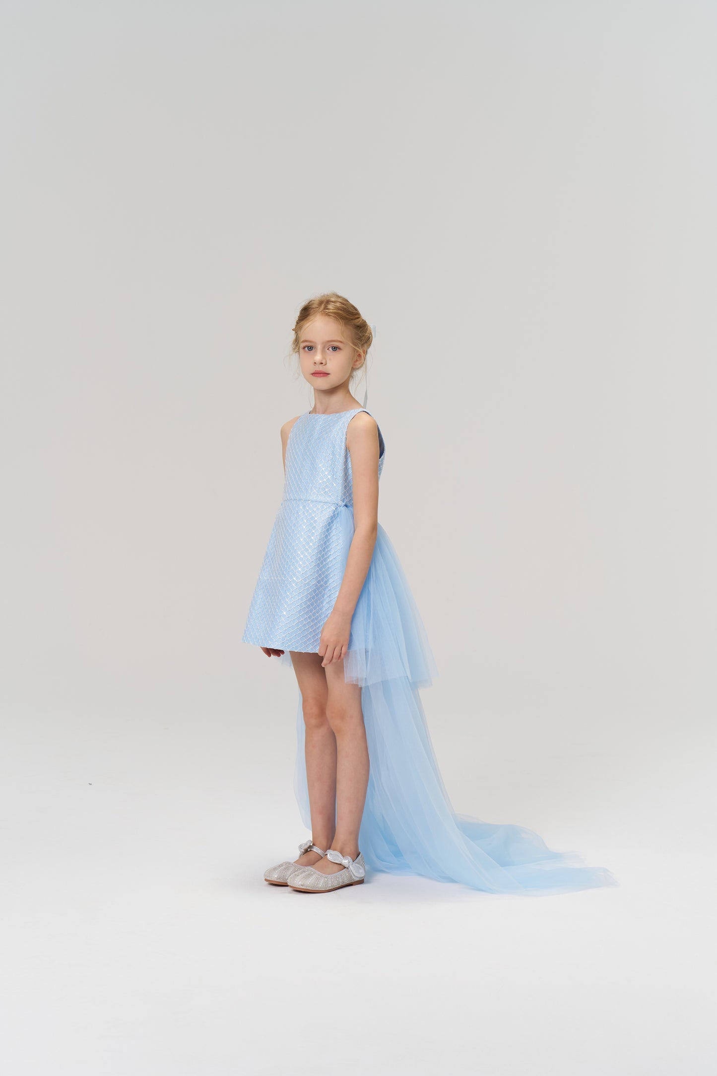 A young girl models the KIKI and JOJO Radiance Sequin Overlay Dress, featuring a light blue hue and a long sequin tulle train, as she poses against a plain white background.