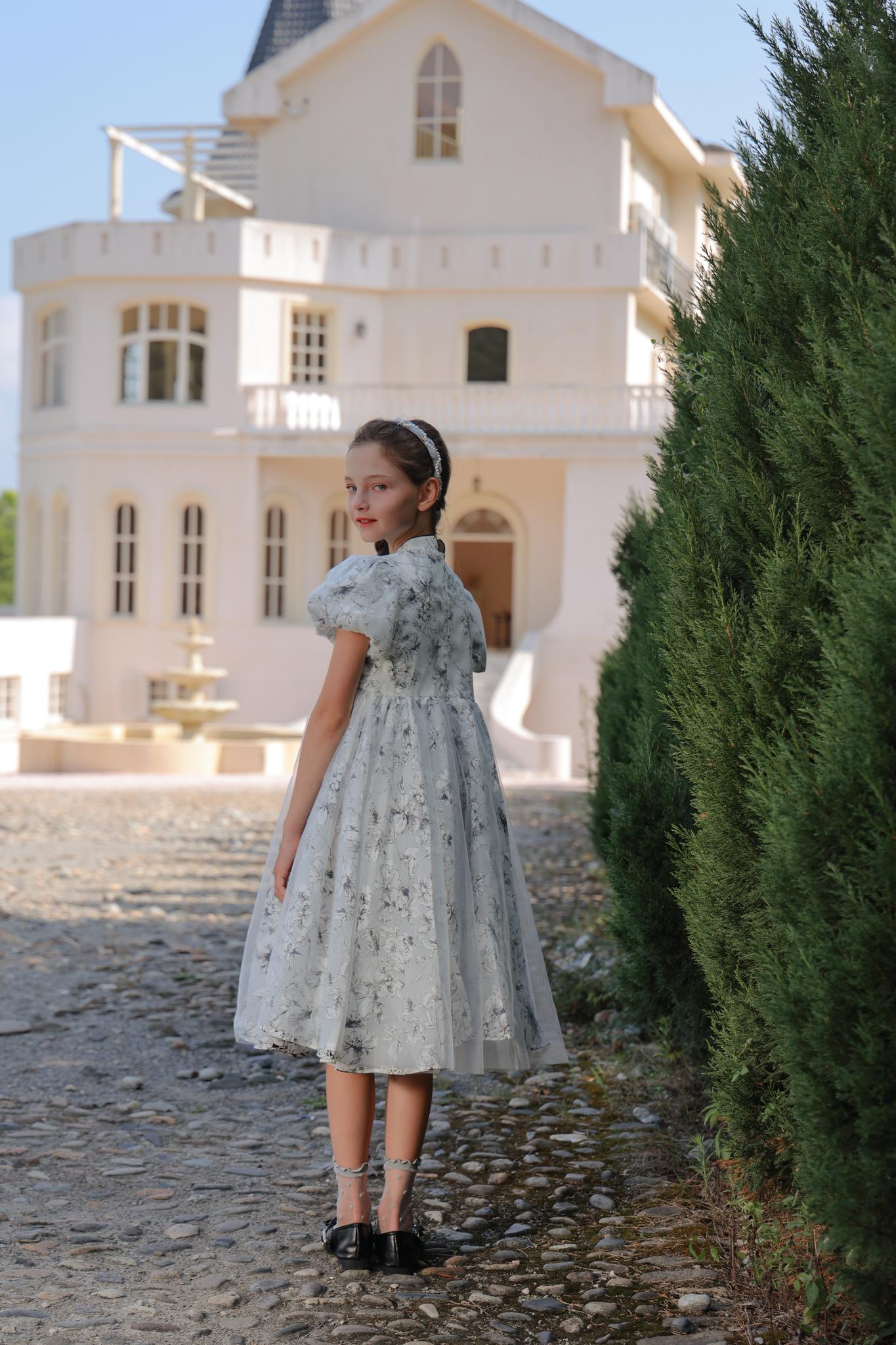 A young girl wears the KIKI and JOJO Girls Cheongsam Dress – Ethereal Blossom Puff Sleeve Tulle, standing on a cobblestone path and looking back, with a large white house and tall greenery behind her.