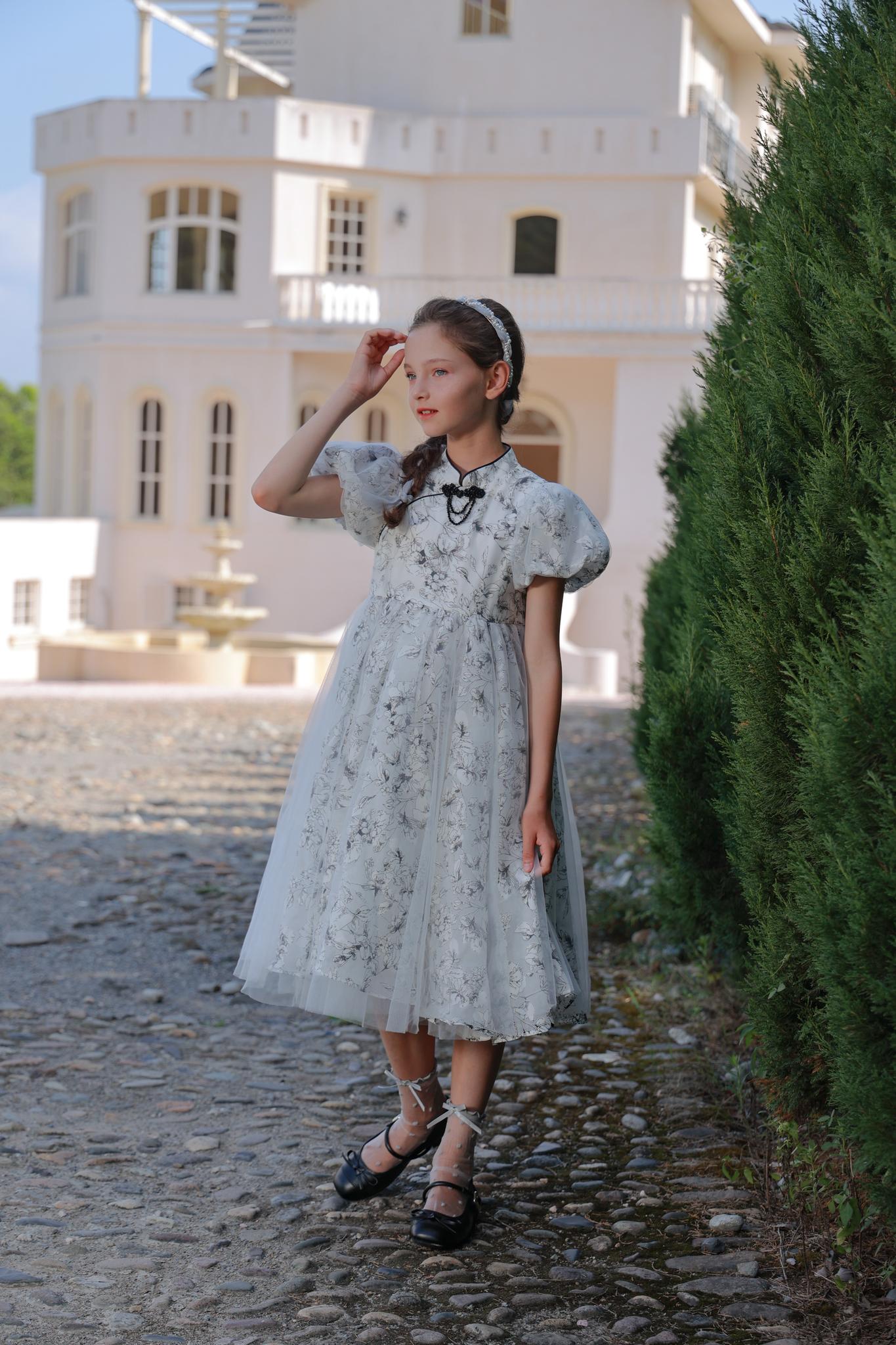 A girl wears the KIKI and JOJO Girls Cheongsam Dress – Ethereal Blossom Puff Sleeve Tulle with black shoes, standing on a cobblestone path by a tall green hedge and a large white building in the background.