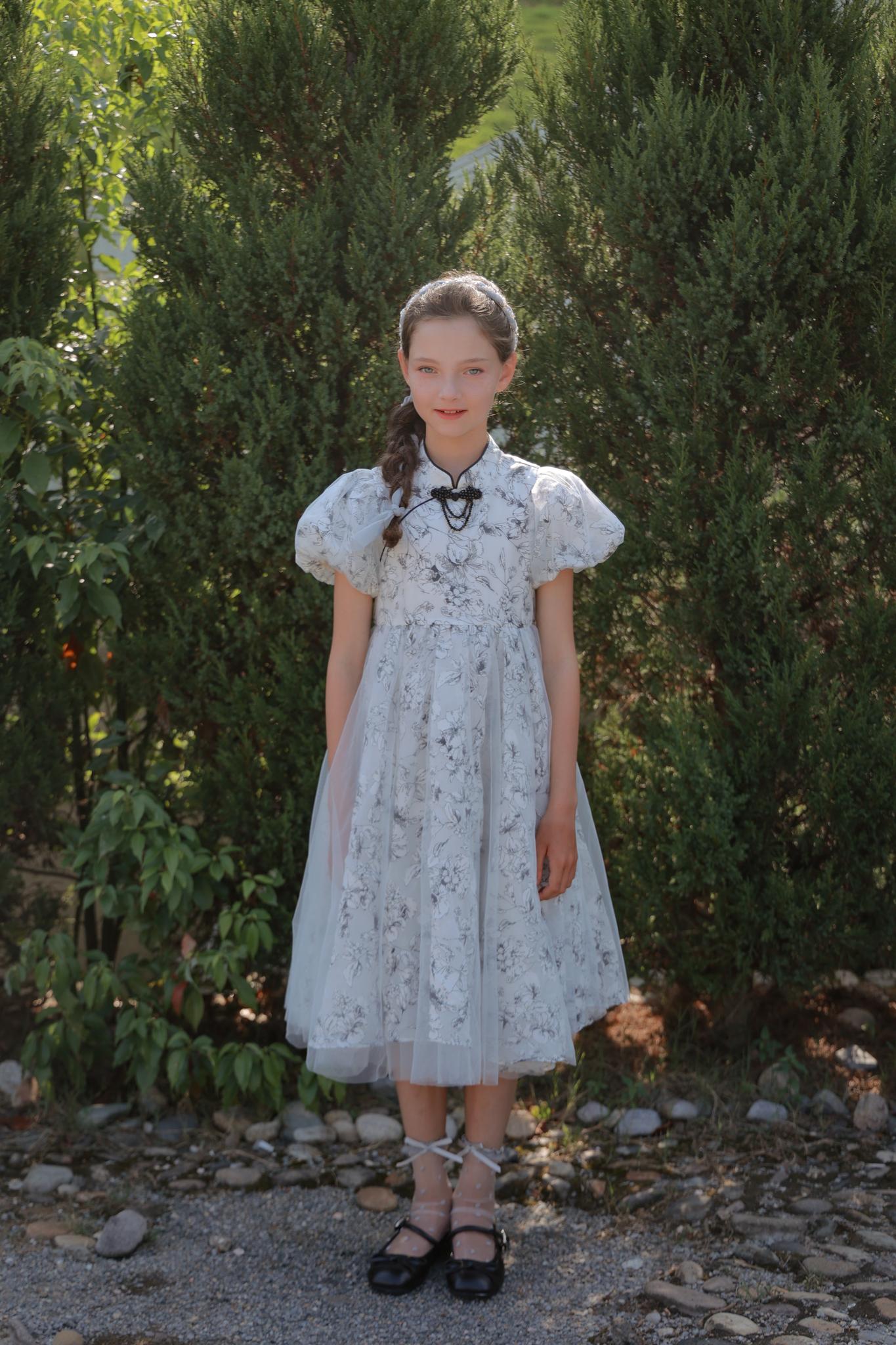 A young girl faces the camera outdoors in front of green shrubs, wearing the KIKI and JOJO Girls Cheongsam Dress – Ethereal Blossom Puff Sleeve Tulle.