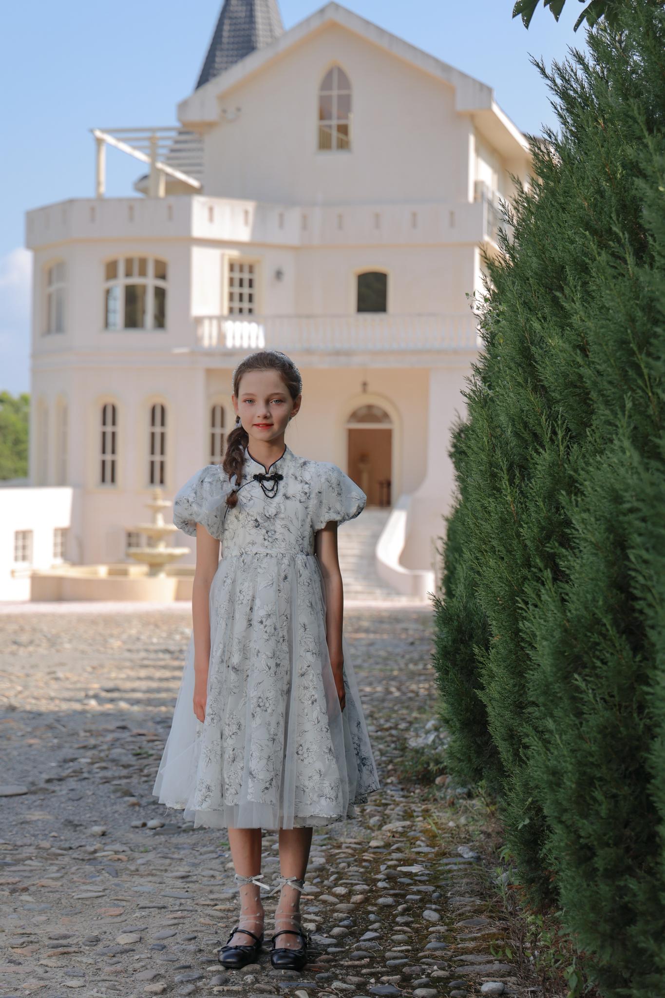 A young girl in the KIKI and JOJO Girls Cheongsam Dress – Ethereal Blossom Puff Sleeve Tulle stands on a cobblestone path before a white house with tall windows and a pointed roof.