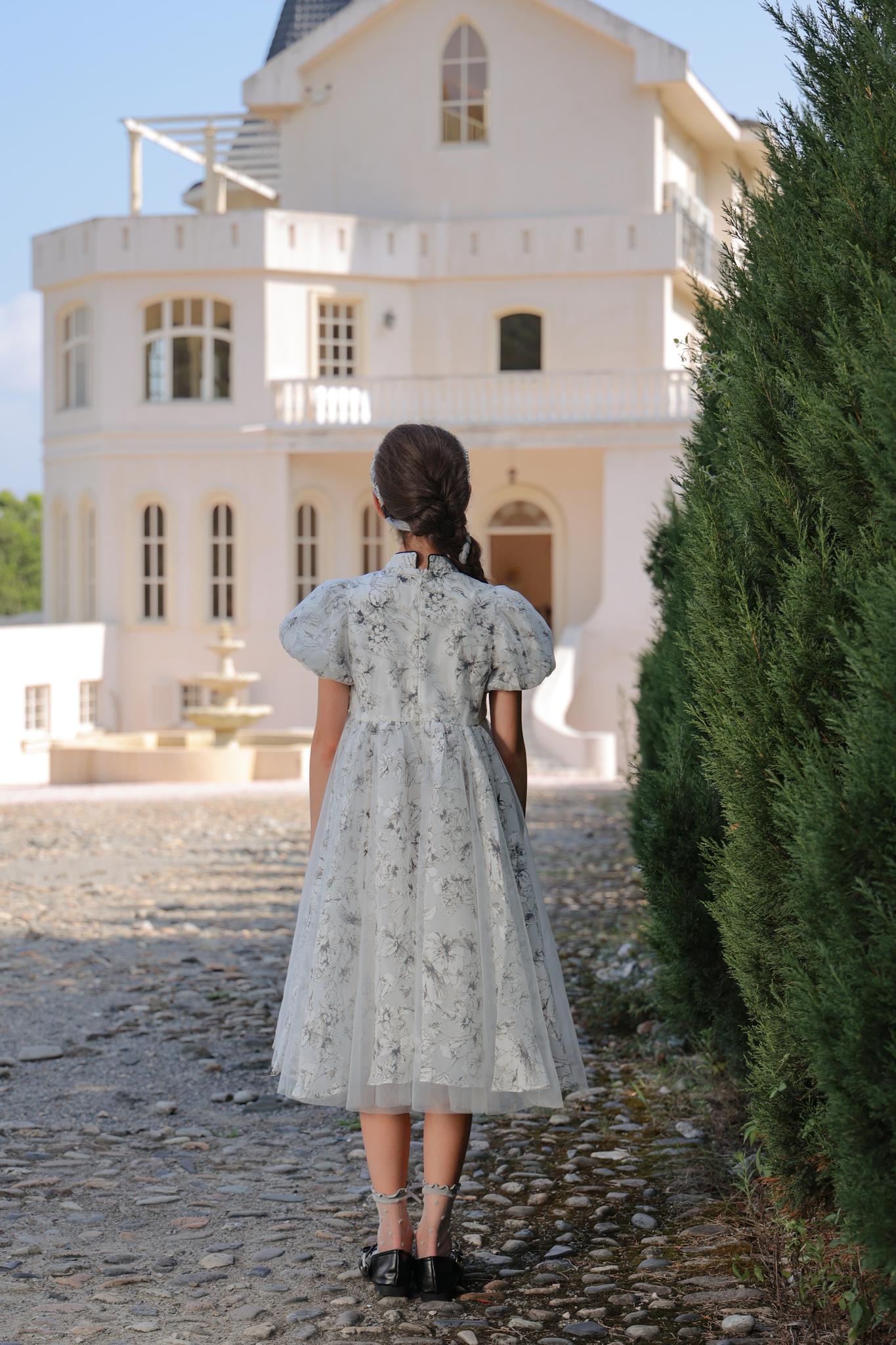 A girl wearing the KIKI and JOJO Girls Cheongsam Dress – Ethereal Blossom Puff Sleeve Tulle stands on a cobblestone path facing a large white house, with trees to the right.