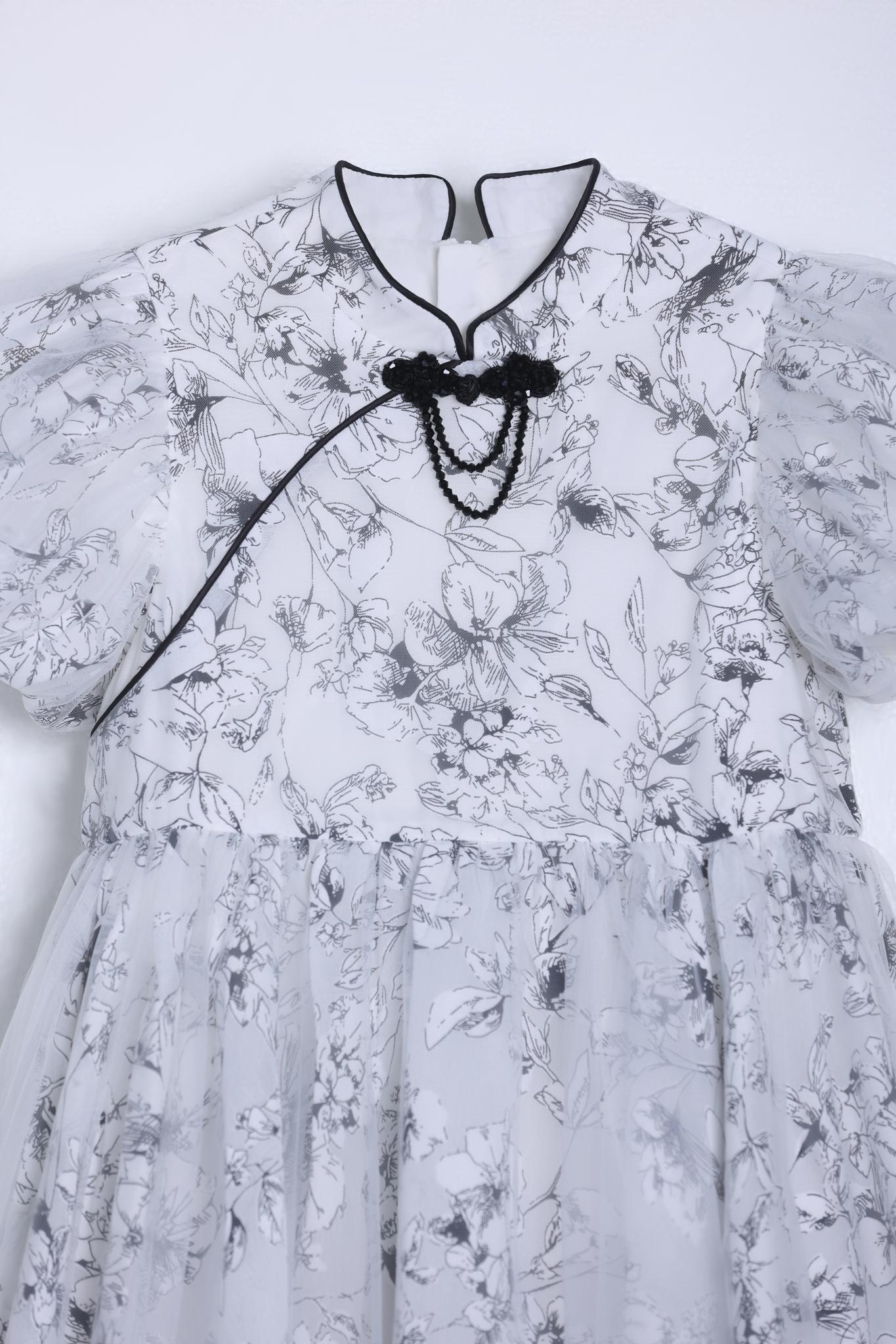 The KIKI and JOJO Girls Cheongsam Dress – Ethereal Blossom Puff Sleeve Tulle features a white base with black floral pattern, puffed short sleeves, tulle overlay skirt, mandarin collar, and a black decorative knot on the chest.