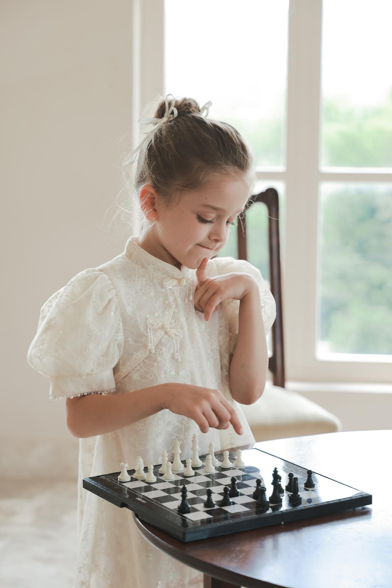 A young girl wearing the KIKI and JOJO Puff Sleeve Tulle Cheongsam Dress with Pearl-Embroidered Cuffs stands by a table, thoughtfully playing chess in a bright room filled with natural light from large windows.
