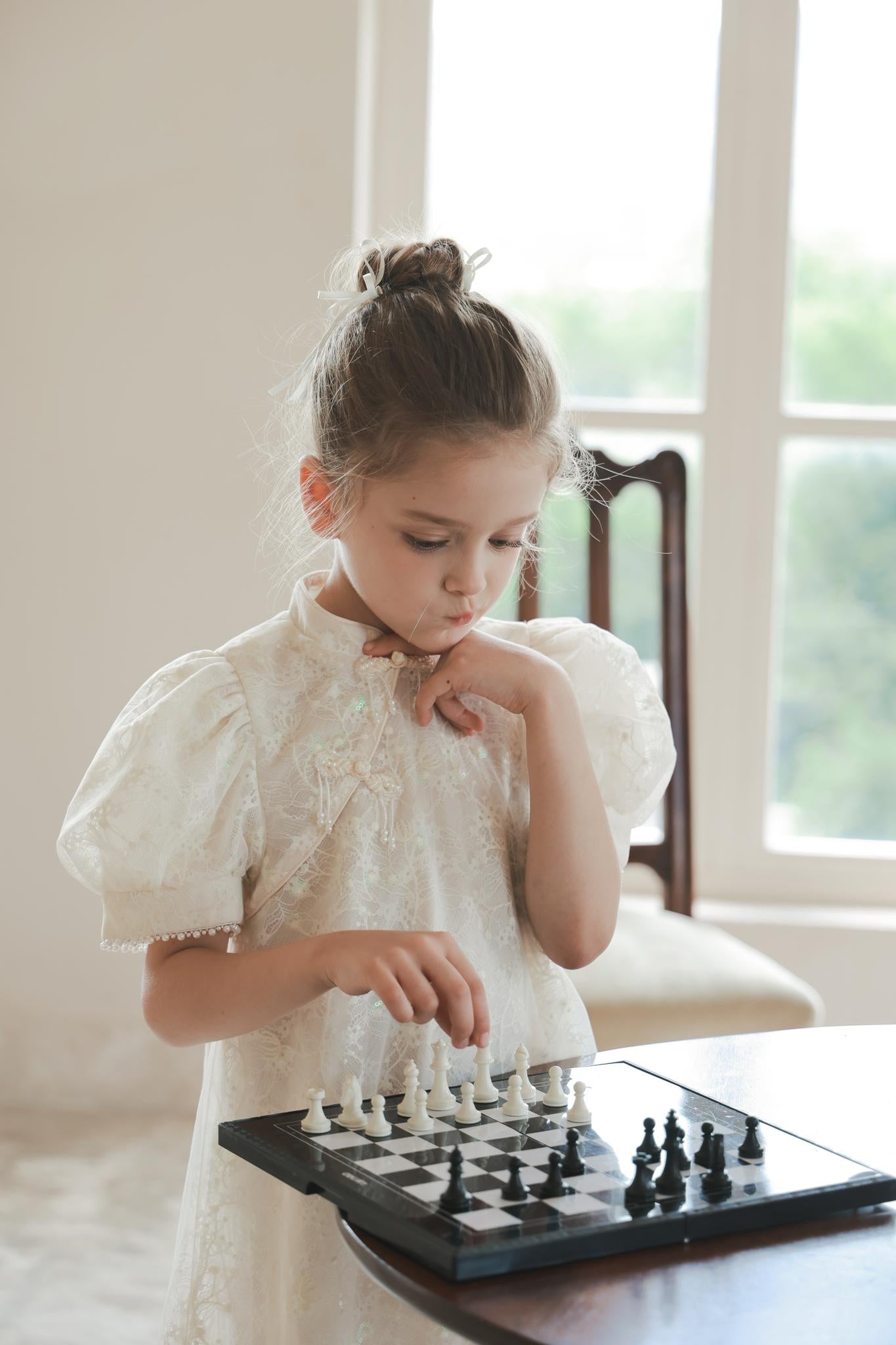 A young girl in the KIKI and JOJO Puff Sleeve Tulle Cheongsam Dress with Pearl-Embroidered Cuffs stands indoors, thoughtfully playing chess at a small board.