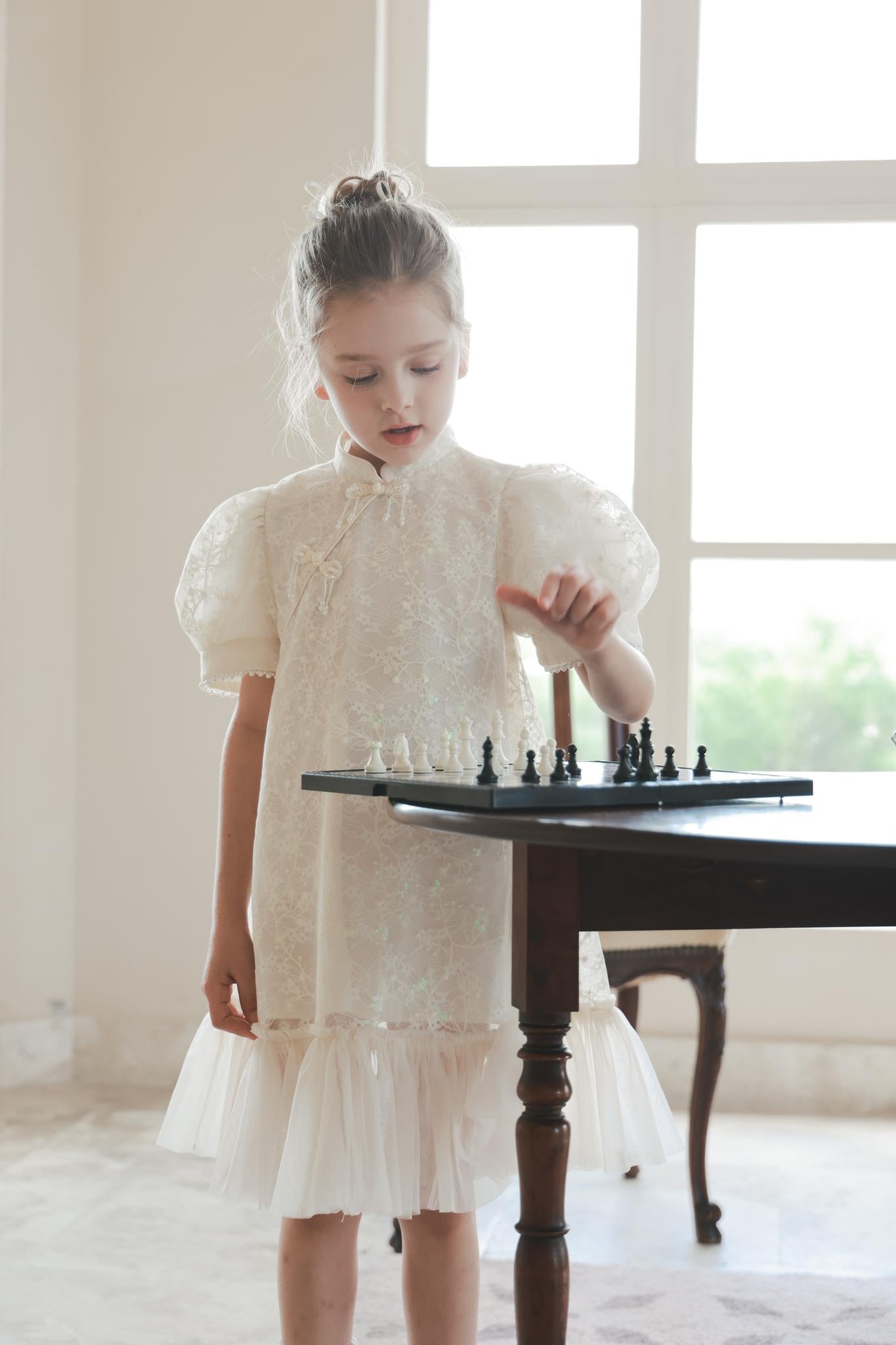 A young girl in the KIKI and JOJO Puff Sleeve Tulle Cheongsam Dress with Pearl-Embroidered Cuffs plays chess at a sunlit table near a large window.