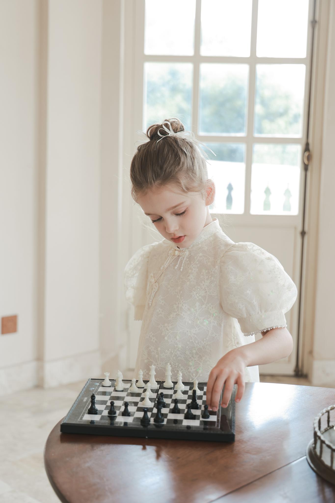 A young girl wearing the KIKI and JOJO Puff Sleeve Tulle Cheongsam Dress with Pearl-Embroidered Cuffs plays chess at a wooden table in a bright room with large windows.