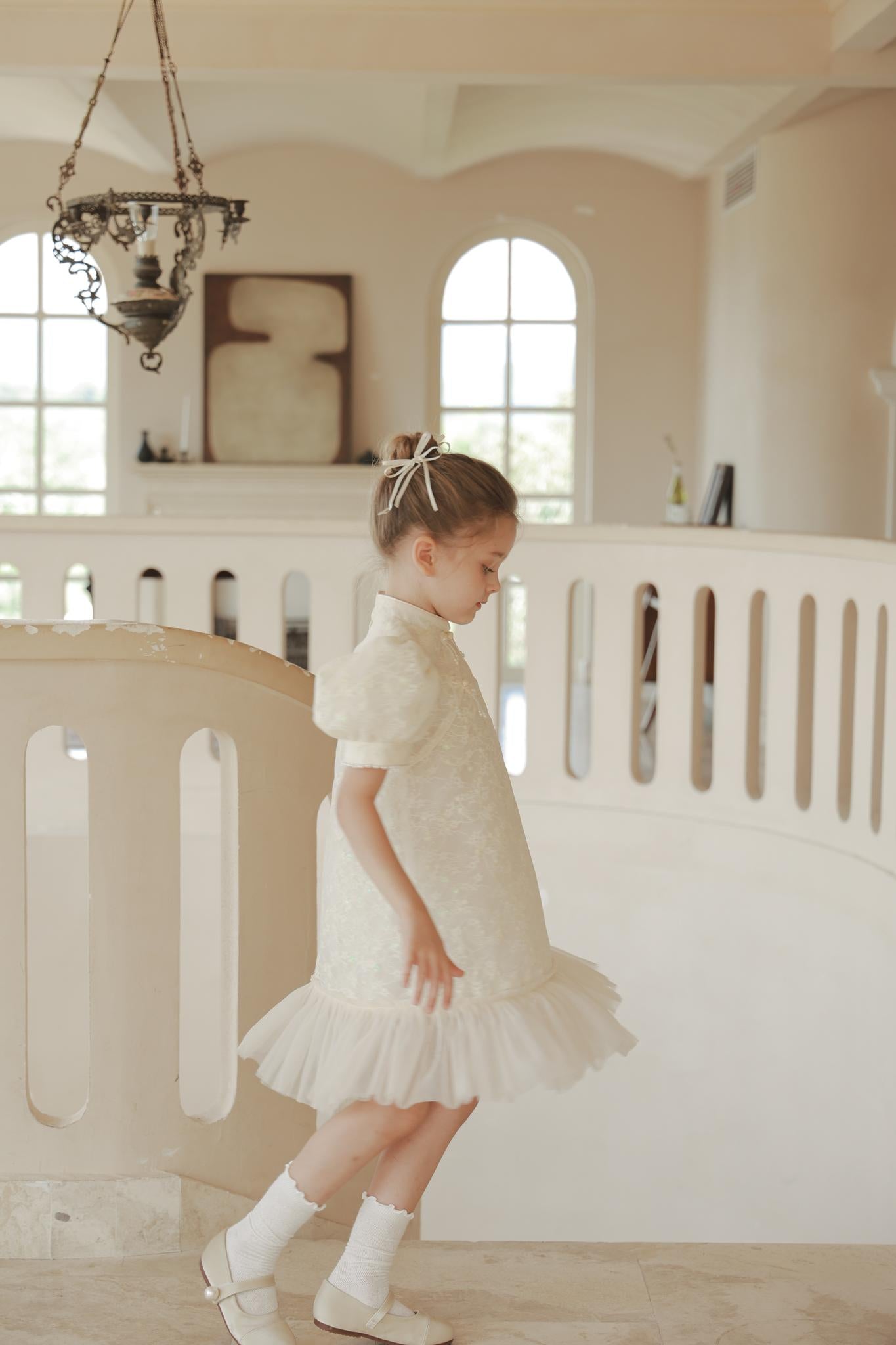 A young girl in the KIKI and JOJO Puff Sleeve Tulle Cheongsam Dress with Pearl-Embroidered Cuffs walks in a bright, elegant interior with arched windows, sparkling chandeliers, and a curved railing.
