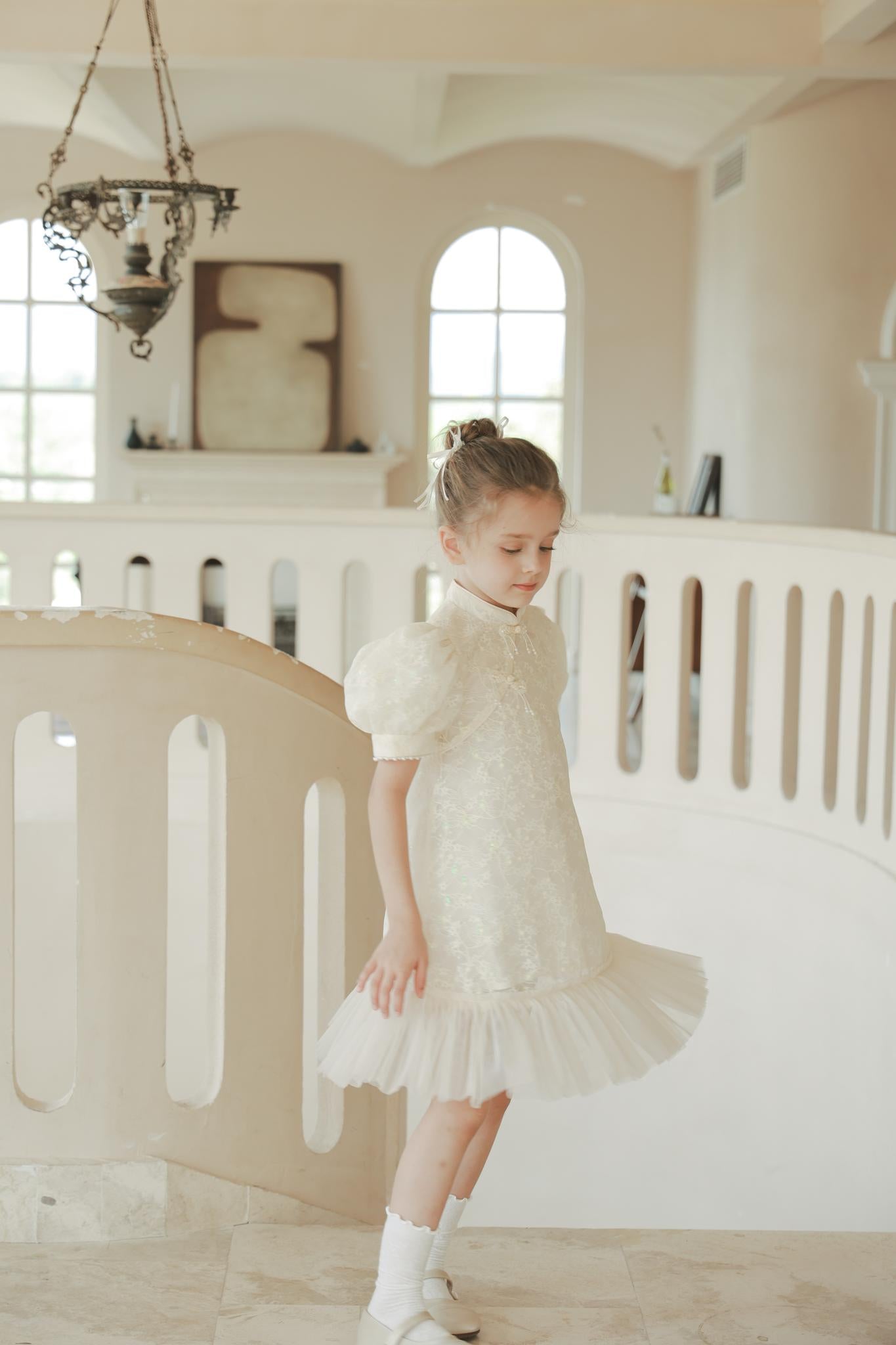 A young girl stands on an indoor staircase, light streaming through arched windows, wearing the KIKI and JOJO Puff Sleeve Tulle Cheongsam Dress with Pearl-Embroidered Cuffs.