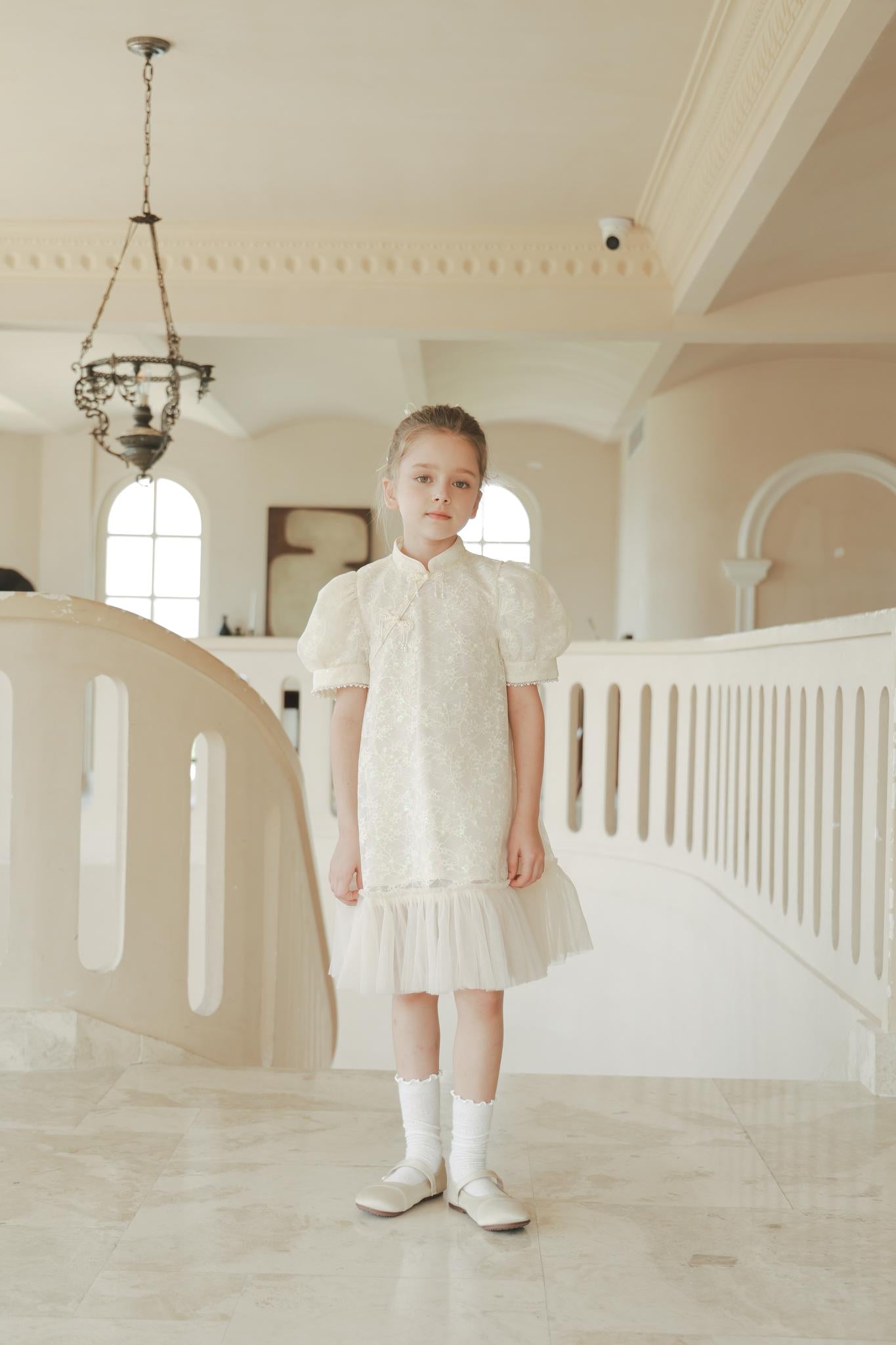 A young girl wears the KIKI and JOJO Puff Sleeve Tulle Cheongsam Dress with Pearl-Embroidered Cuffs, standing indoors on a marble floor near a white railing, with neutral walls and arched windows in the background.