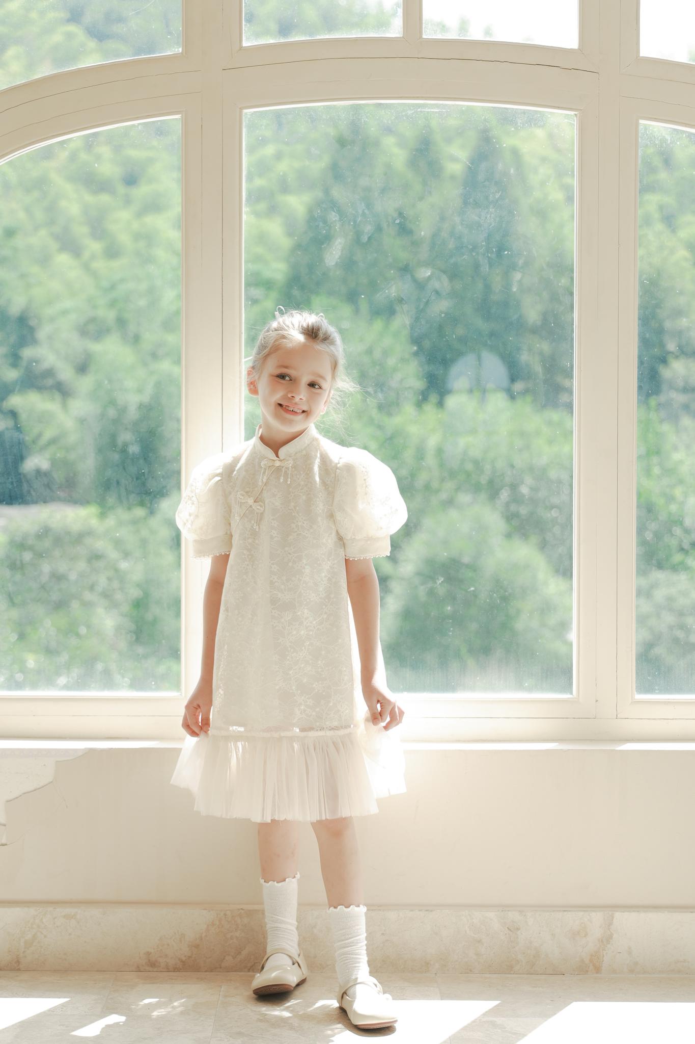 A young girl wears the KIKI and JOJO Puff Sleeve Tulle Cheongsam Dress with Pearl-Embroidered Cuffs, standing before a large window overlooking a green, sunlit landscape.