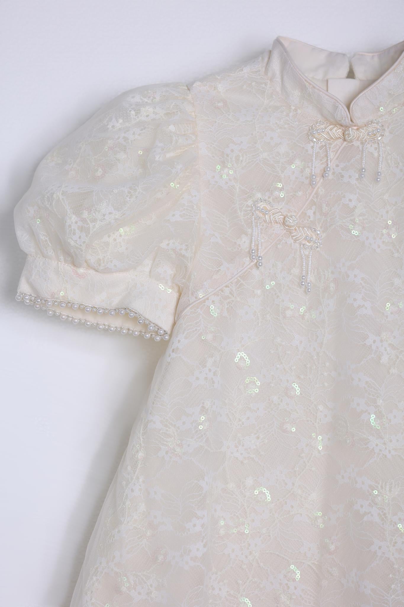 Close-up of the KIKI and JOJO Puff Sleeve Tulle Cheongsam Dress, showcasing pearl-embroidered cuffs, decorative knots, and delicate sequins on an off-white lace overlay.
