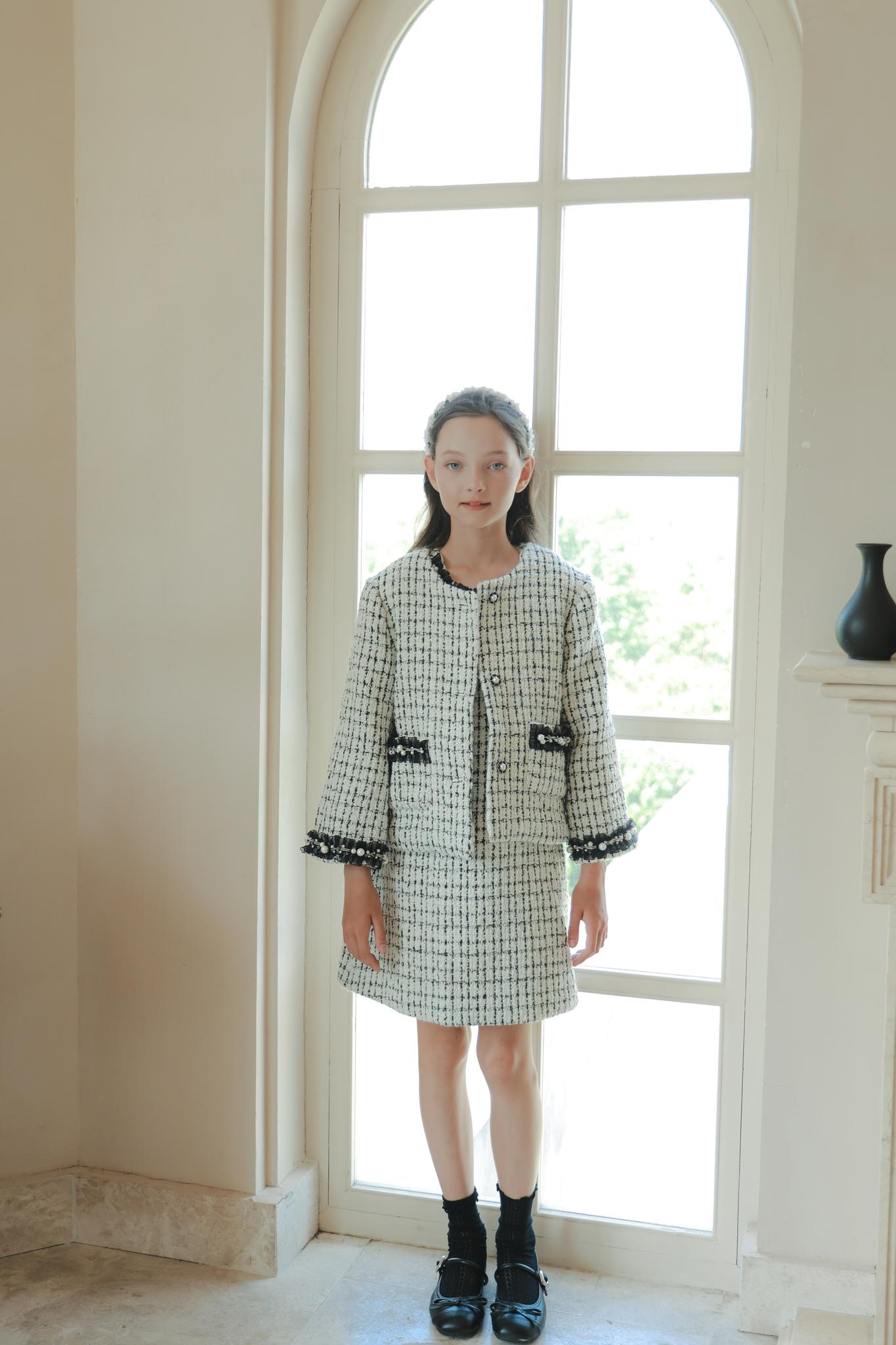 A young woman stands indoors by a tall arched window, wearing the KIKI and JOJO Girls Tweed Jacket – Black & White Checkered Pearl with a matching skirt, paired with black shoes and socks.