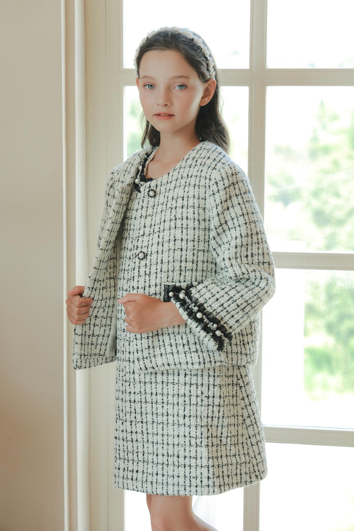 A young girl stands indoors by a window, wearing the KIKI and JOJO Girls Tweed Jacket – Black & White Checkered Pearl, paired with a matching skirt and headband.