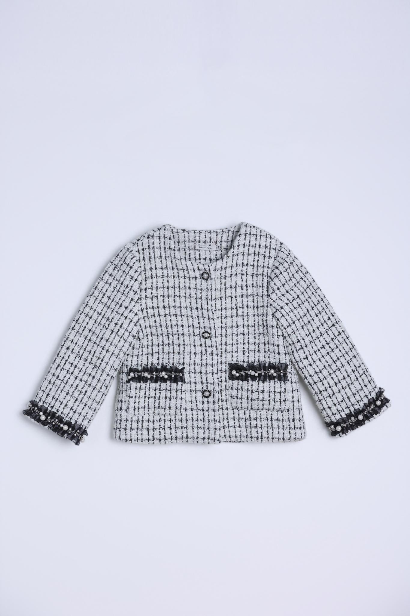 The KIKI and JOJO Girls Tweed Jacket in Black & White Checkered Pearl features decorative black fringe on the pockets and cuffs, classic black buttons, and is displayed laid flat against a plain light background.