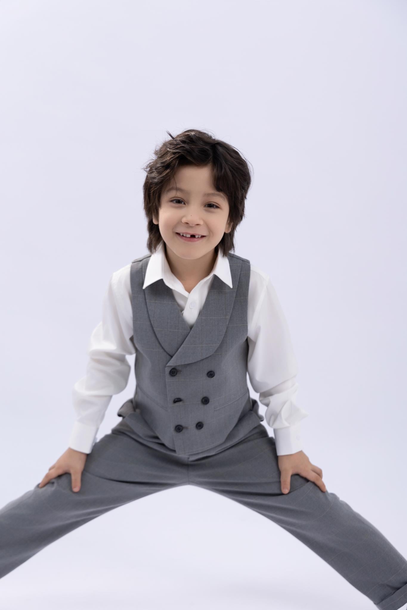 A boy wears the KIKI and JOJO Kids Suit Jacket in light gray checkered, wrinkle-free fabric, posing with legs apart and hands on his knees against a plain light background. Suitable for ages 3–14 years.