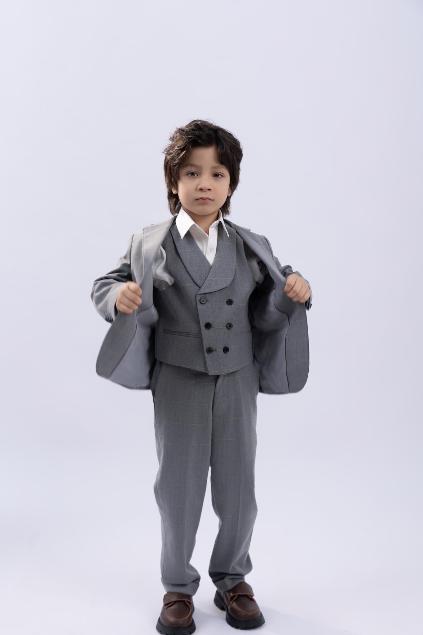 A young boy stands facing forward against a plain light background, wearing KIKI and JOJO’s checkered Kids Suit Pants in light gray (3-14 years, adjustable), paired with a gray suit and brown shoes, holding open the sides of his jacket.