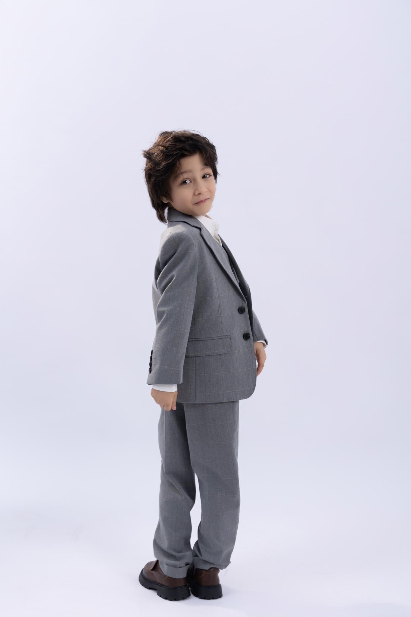 A young boy stands against a plain light background, looking over his shoulder and wearing the KIKI and JOJO Kids Suit Jacket in light gray checkered wrinkle-free fabric (ages 3-14).
