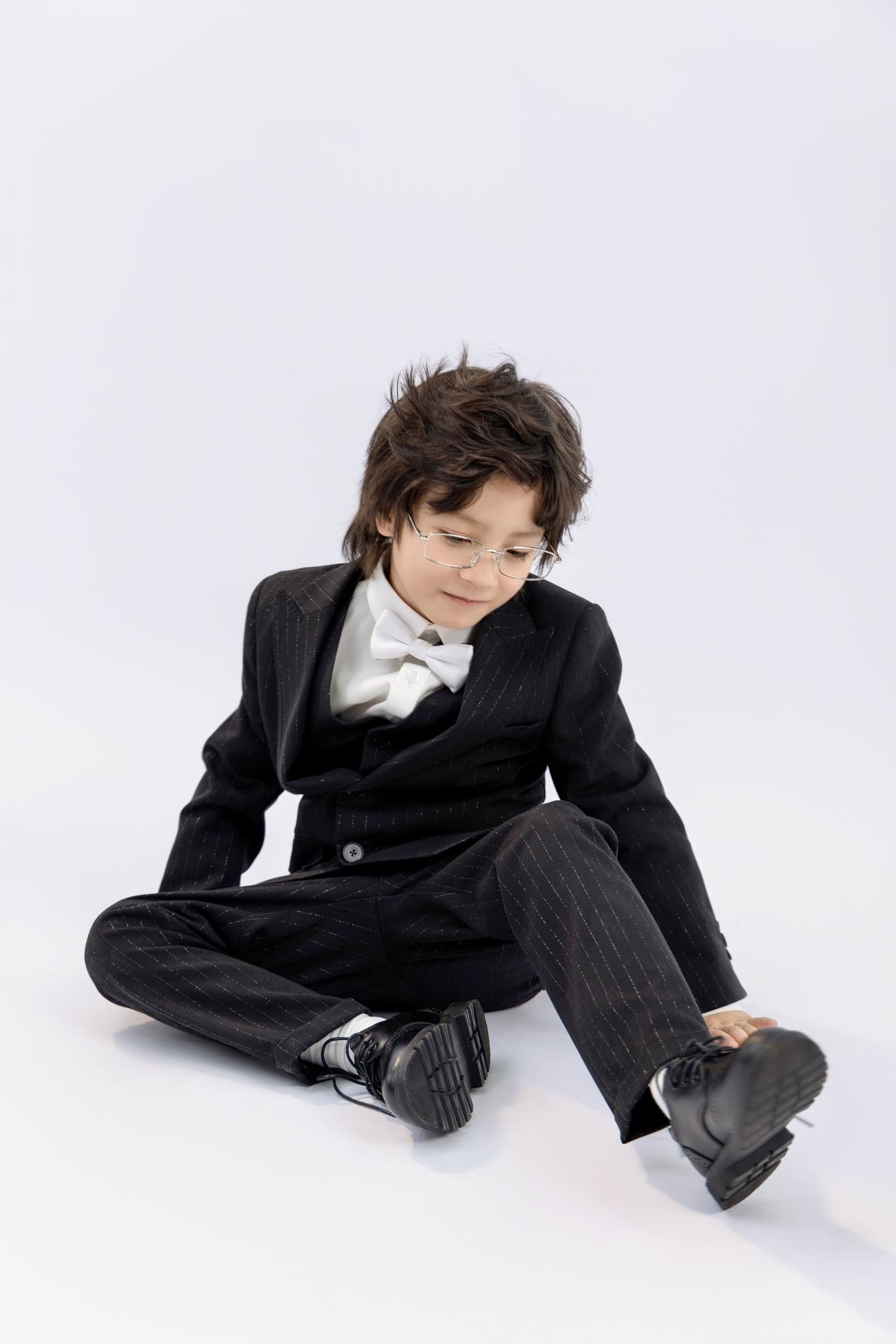 A young boy wearing KIKI and JOJO's Kids Suit Pants – Black (Classic Fit, Wrinkle-Free, ages 3-14) with a white bow tie sits on the floor and touches his shoe against a plain white background.