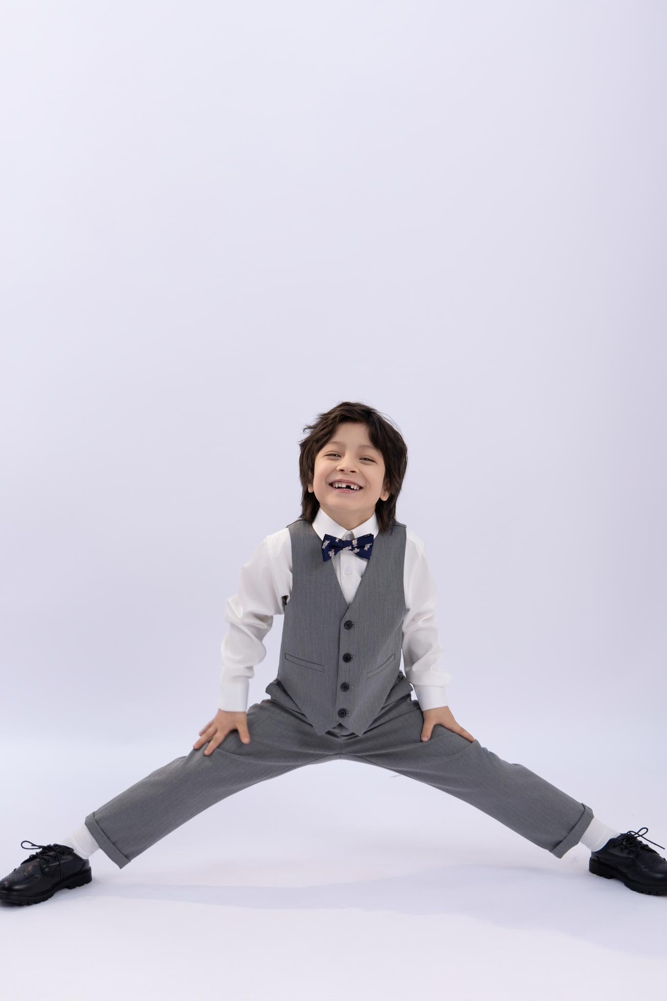 A young boy in a gray suit and bow tie does a split, smiling against a white background—highlighting the classic fit and wrinkle-free fabric of KIKI and JOJO Kids Suit Pants – Gray (3-14 Years).