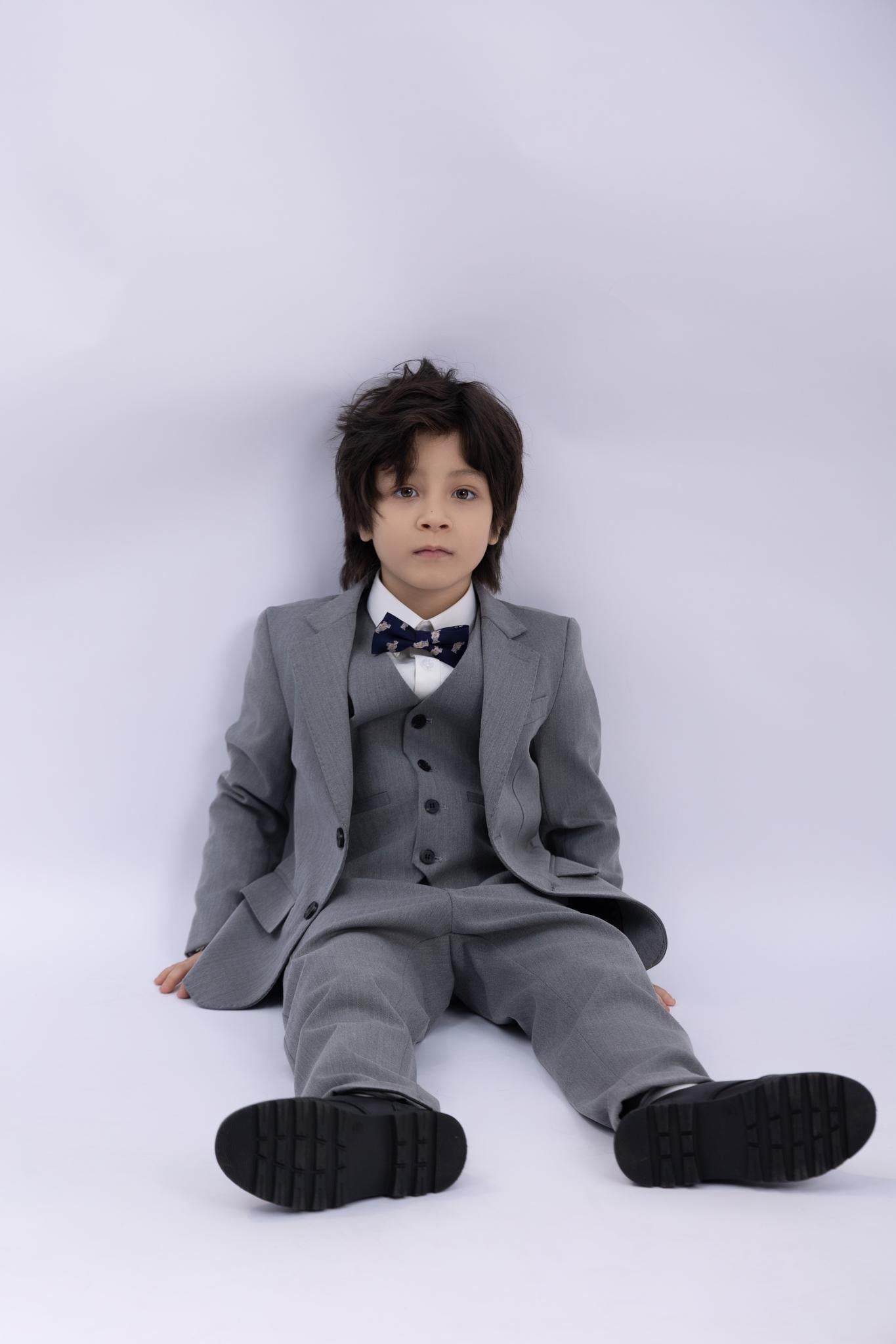 A young boy sits on the floor against a white background wearing a KIKI and JOJO Kids Suit Jacket – Gray (3-14 Years), Classic Fit, Wrinkle-Free, paired with a bow tie.