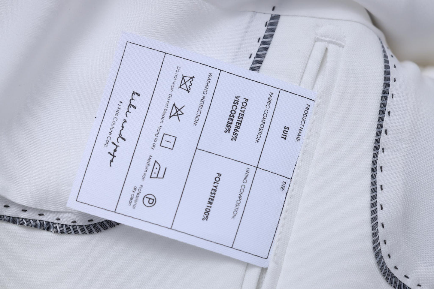 Close-up of the KIKI and JOJO Kids Suit Jacket – White (3-14 Years), Classic Fit, wrinkle-free, with a label showing care instructions, material details, and a signature for a timeless look.