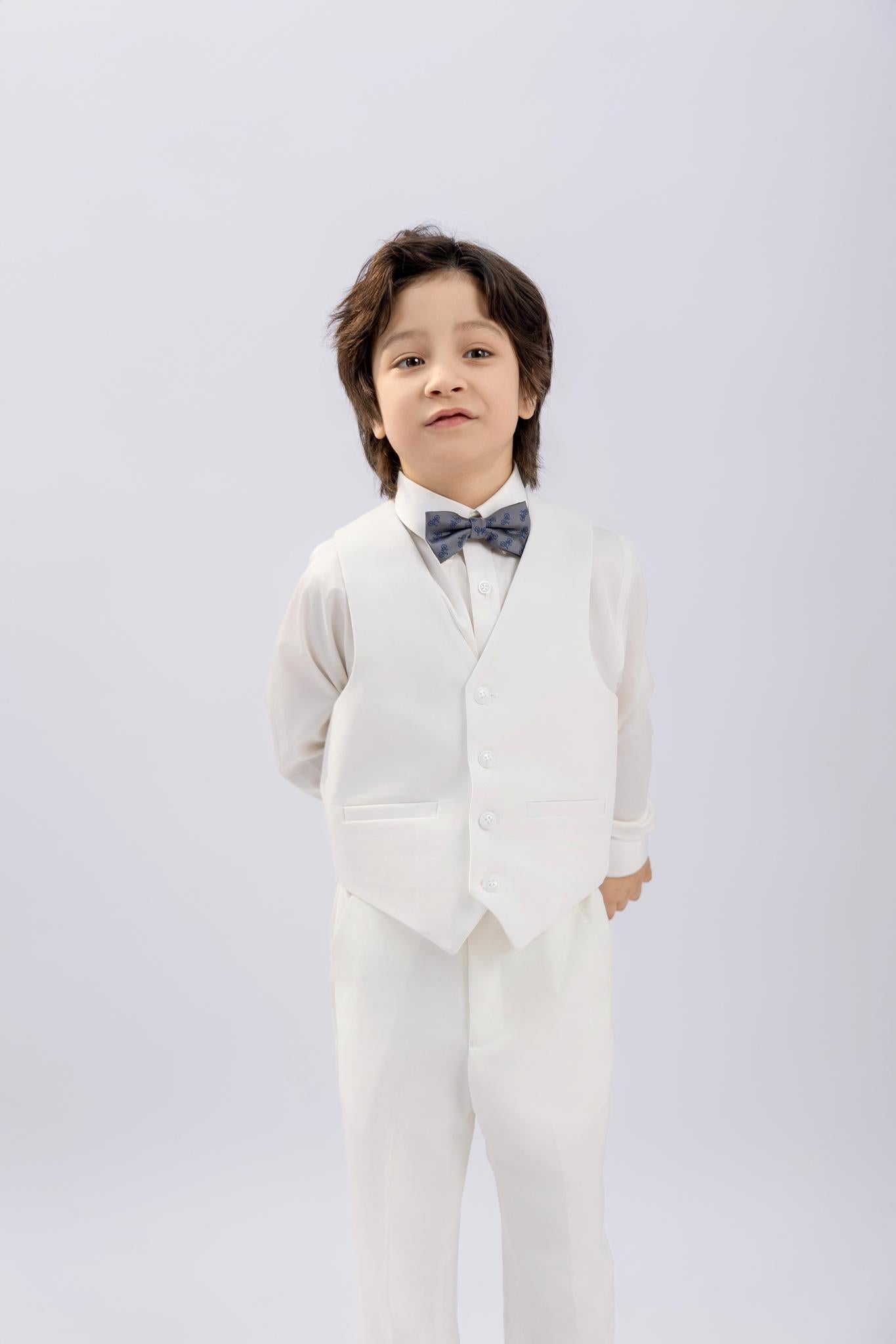 A young boy stands against a plain background, hands behind his back, dressed in a KIKI and JOJO Kids Suit Vest – White (3-14 Years), a classic fit and wrinkle-free design, paired with a white shirt and gray bow tie.