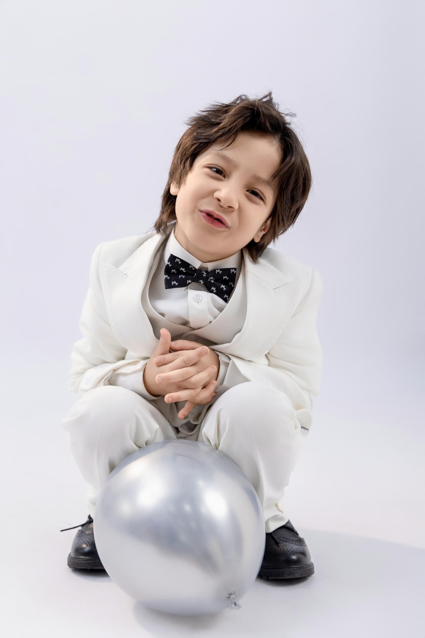A young child in a KIKI and JOJO Kids Suit Vest – White (3-14 Years), Classic Fit, Wrinkle-Free, with a black bow tie, sits crouched on the floor beside a silver balloon against a plain light background.