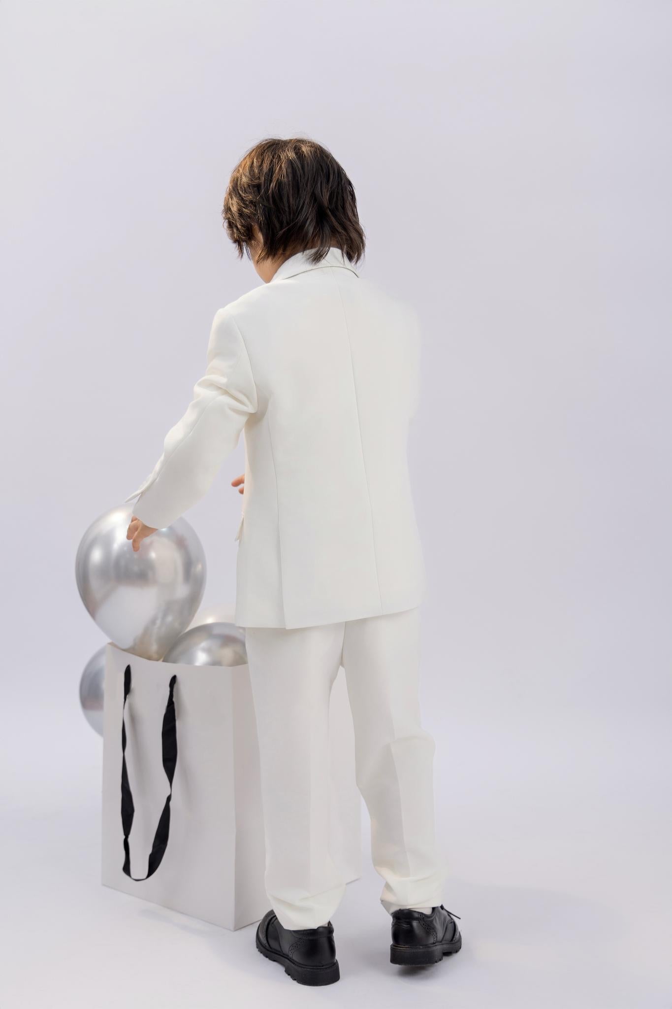 A child wearing the KIKI and JOJO Kids Suit Vest – White (Classic Fit, Wrinkle-Free, 3-14 Years) stands with their back to the camera, reaching into a white bag of silver balloons against a plain background.