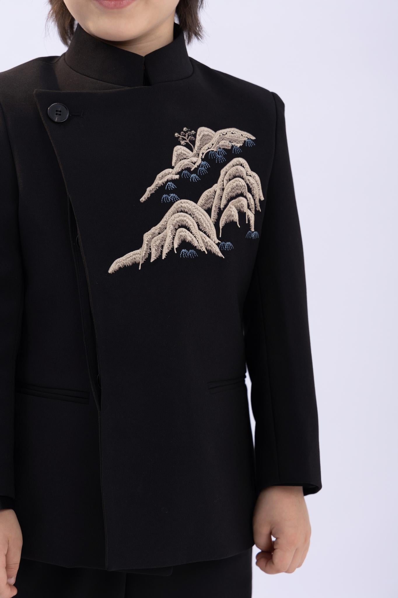 A child wearing the KIKI and JOJO Kids Suit Jacket – Black (Slim-Fit, Wrinkle Free, 3-14 Years) with a classic Mandarin collar and embroidered mountain detail, stands against a plain light background.
