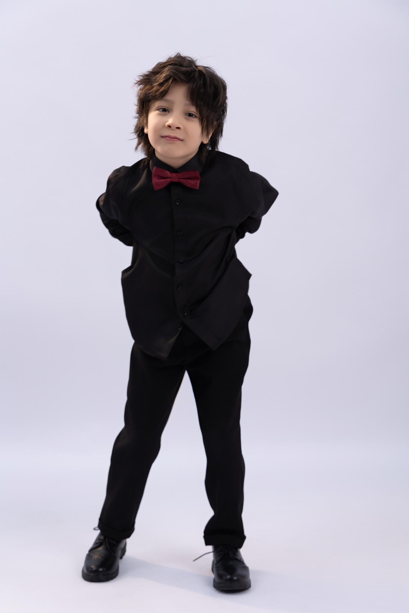 A young boy stands with his hands behind his back, wearing the KIKI and JOJO Kids Dress Shirt – Black Long Sleeve (Classic Fit, 3-14 Years) with black pants, black shoes, and a red bow tie against a plain light background.