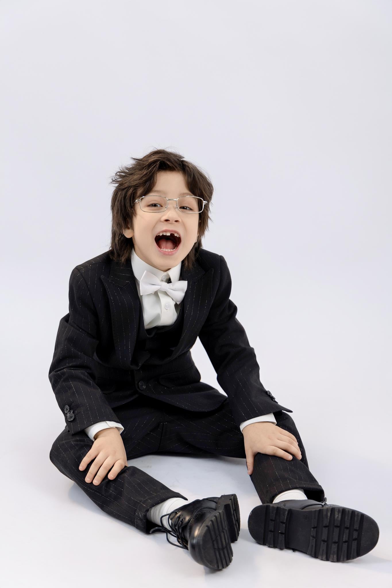A young boy smiles with mouth open while sitting on the floor in a KIKI and JOJO Kids Dress Shirt – White Long Sleeve (3-14 Years), classic fit, paired with a black suit and bow tie, against a plain white background.