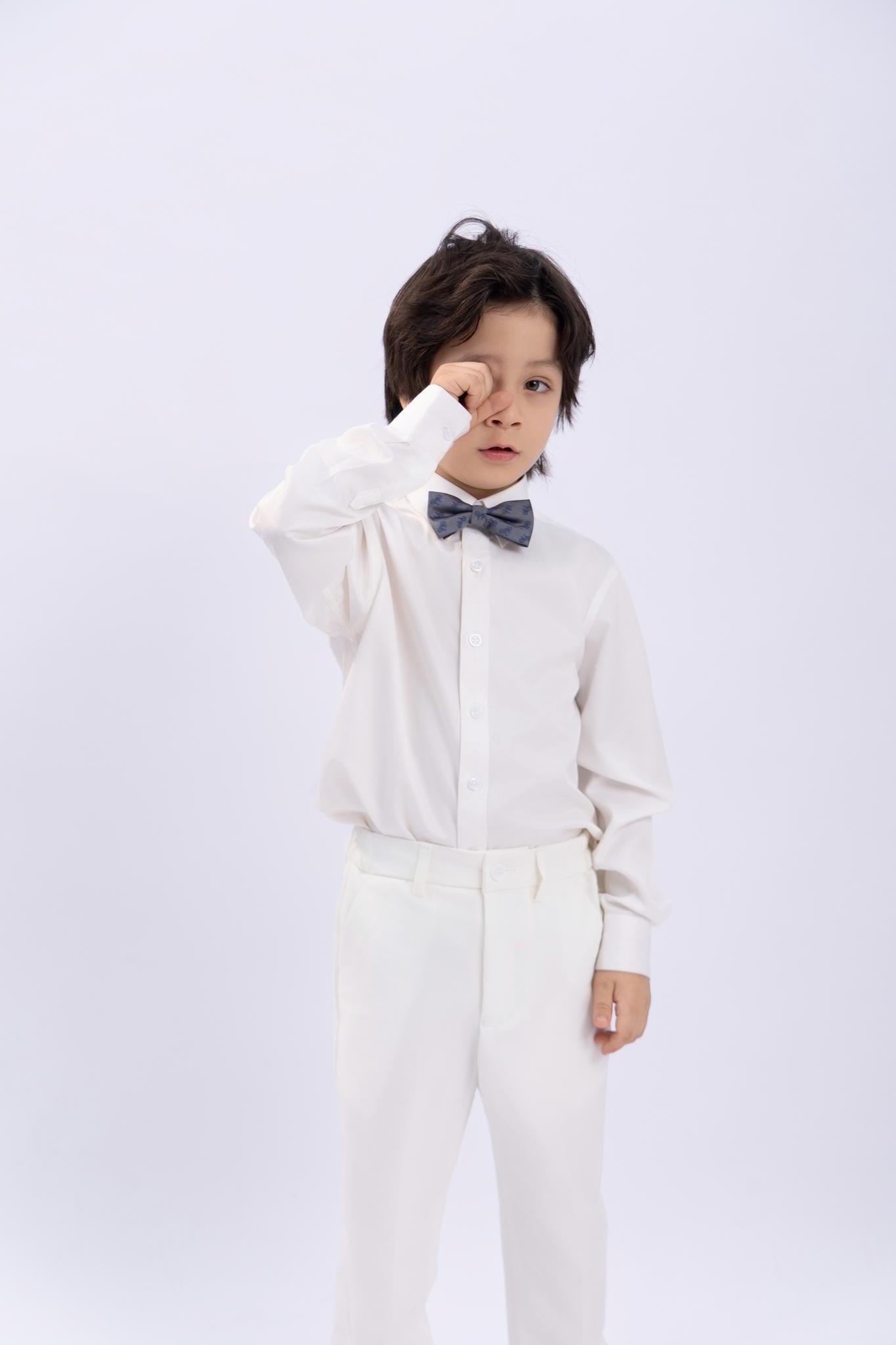 A young boy wearing a white shirt and pants is seen touching his face near his eye, styled with a KIKI and JOJO Kids Bow Tie – Light Gray Bicycle, pre-tied satin and adjustable, standing against a plain light background.