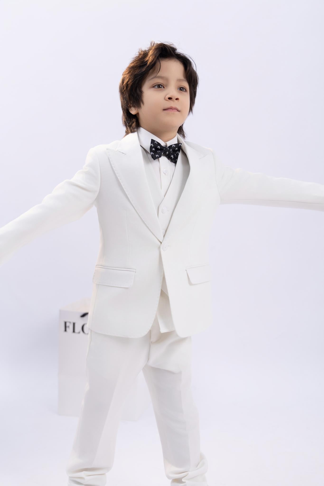 A young boy, arms outstretched against a plain light background, wears a white suit and the KIKI and JOJO Kids Bow Tie – Black Puppy – Pre-Tied Satin – Adjustable.