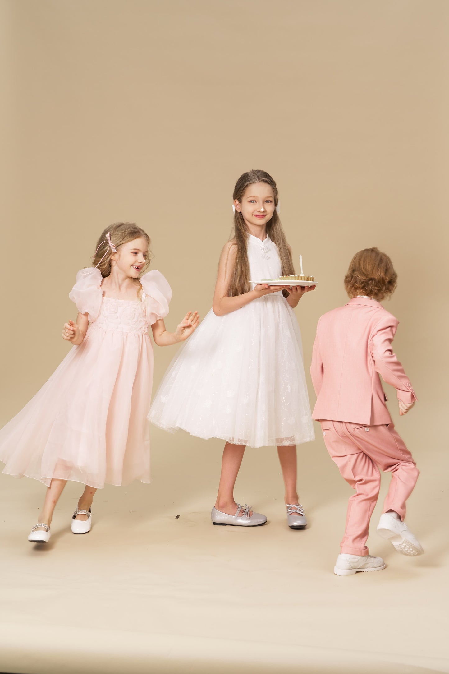 Three children pose against a beige background; one girl wears KIKI and JOJO’s Girls Pink Beaded Bow Hair Clips while holding a cake, another girl smiles, and a boy in a pink suit turns away.