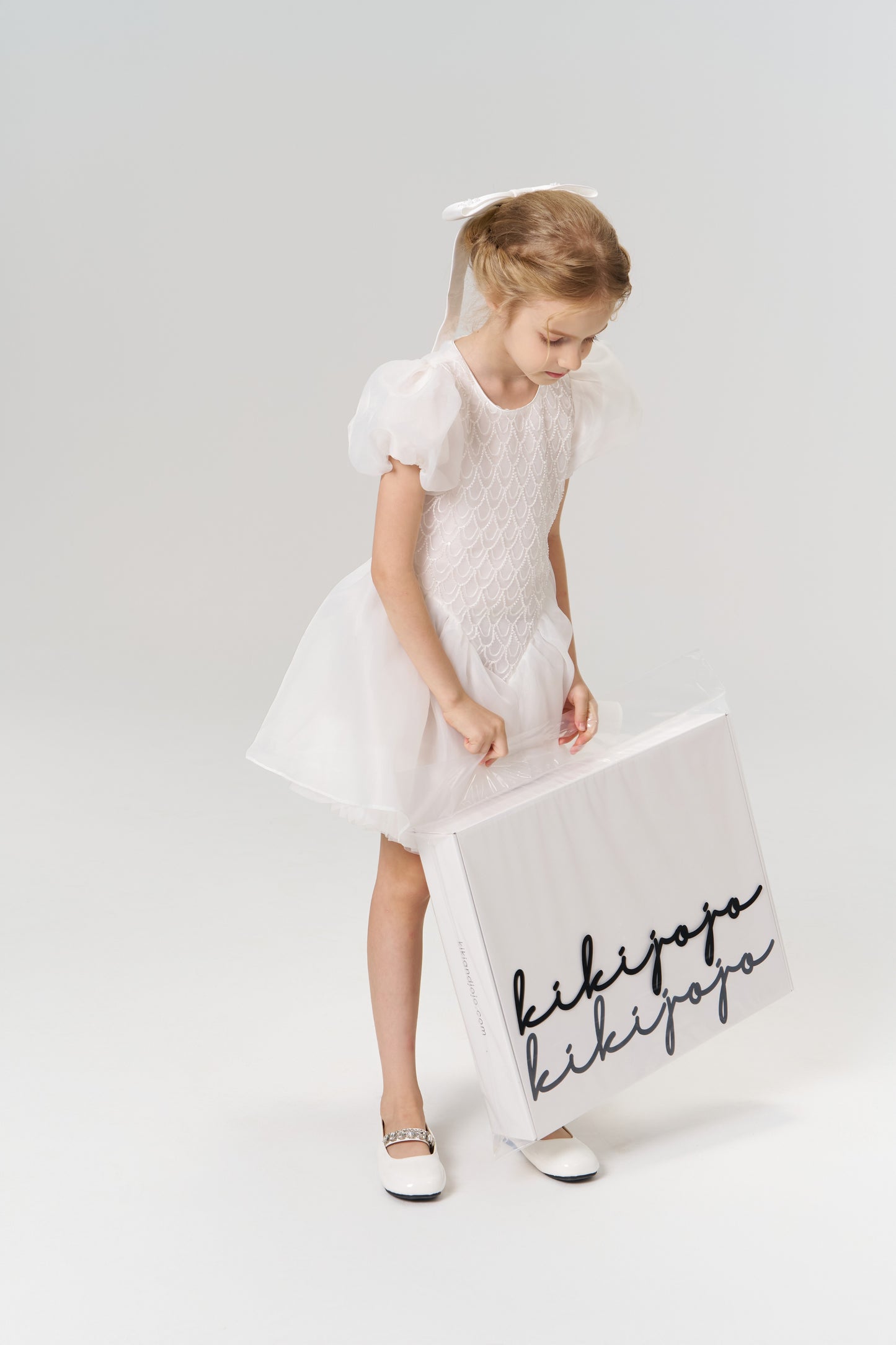 A young flower girl in a white dress with a satin bow wears the Girls Hand-Beaded Pearl Satin Bow Hair Clip by KIKI and JOJO, holding a large white shopping bag with "kikijapa" written in black script.
