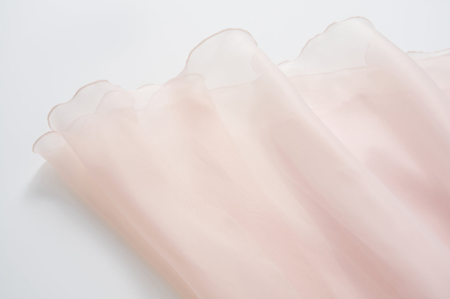 A close-up of the Blush Whisper Dress by KIKI and JOJO shows its sheer, light pink fabric with soft, wavy edges, evoking a princess-style look as it’s spread out on a white surface.