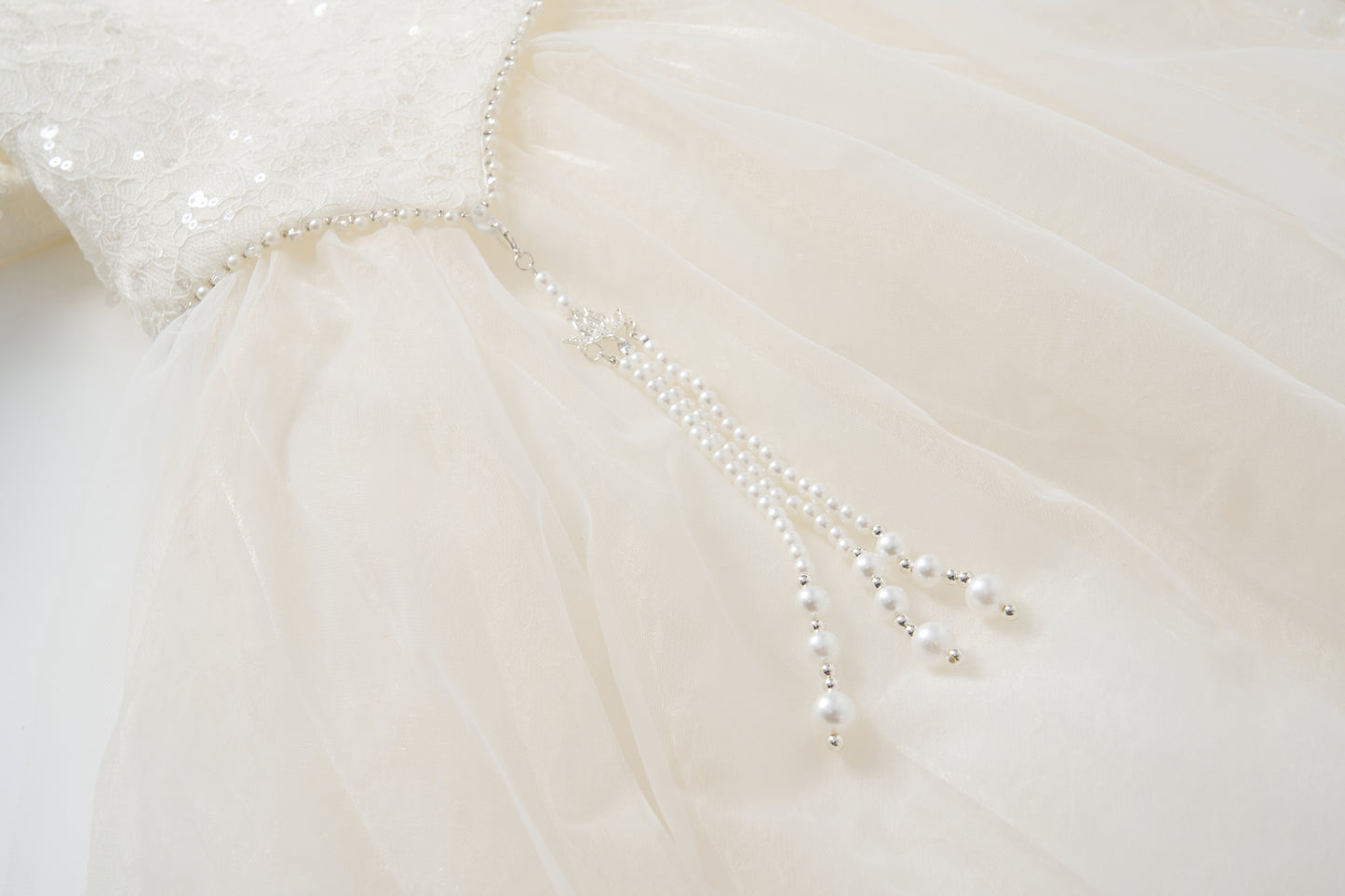 Close-up of the Orchid Whisper Dress by KIKI and JOJO, showcasing a white formal design with a sequin lace bodice and pearl bead strands cascading from the waist for a modern vintage look.