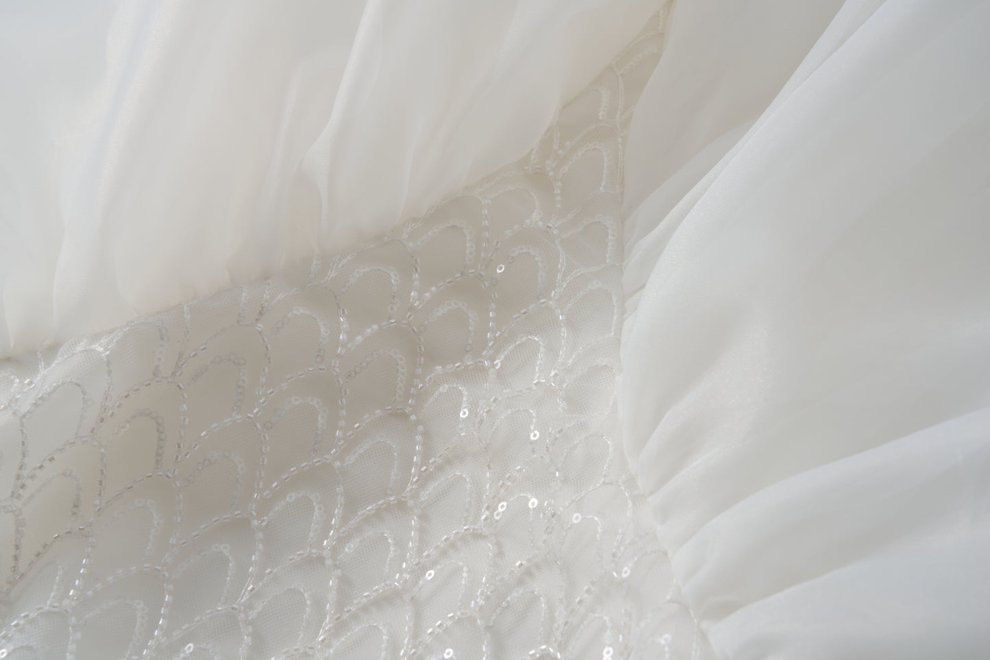 Close-up of the Ballerina Whisper Dress by KIKI and JOJO, featuring white fabric with sheer gathered tulle sides and a central panel adorned with a scalloped sequin pattern, evoking an elegant ballet-inspired design.