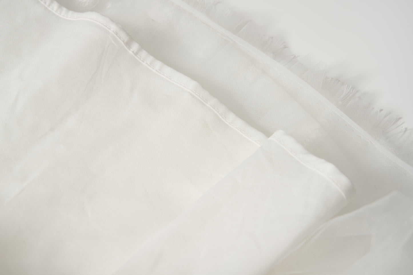 Close-up of the Monochrome Whisper Dress by KIKI and JOJO, showcasing its white, semi-transparent fabric with frayed edge—evoking delicate tulle layers—set against a light background.