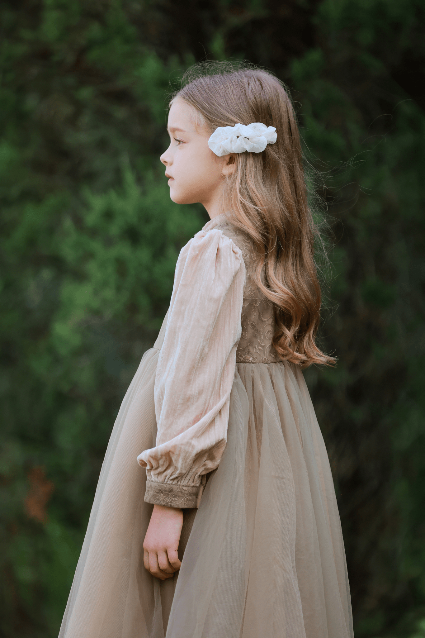 A young girl with long, wavy hair stands in profile, wearing a beige dress and the Girls Chiffon Hair Clip - Ivory by KIKI and JOJO, set against lush green foliage.